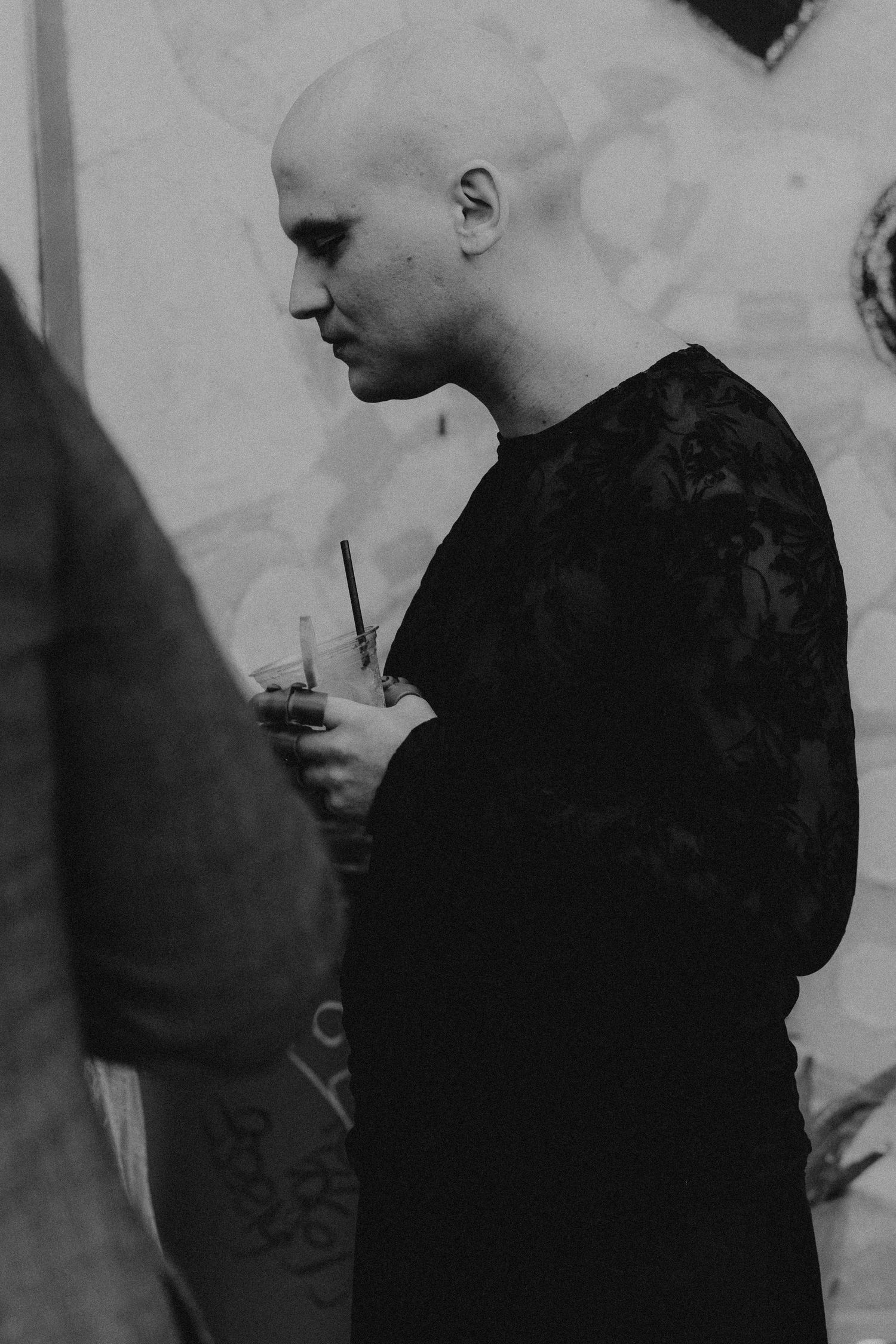 Bald person in black outfit holding a drink with a straw, captured in moody black and white photography.