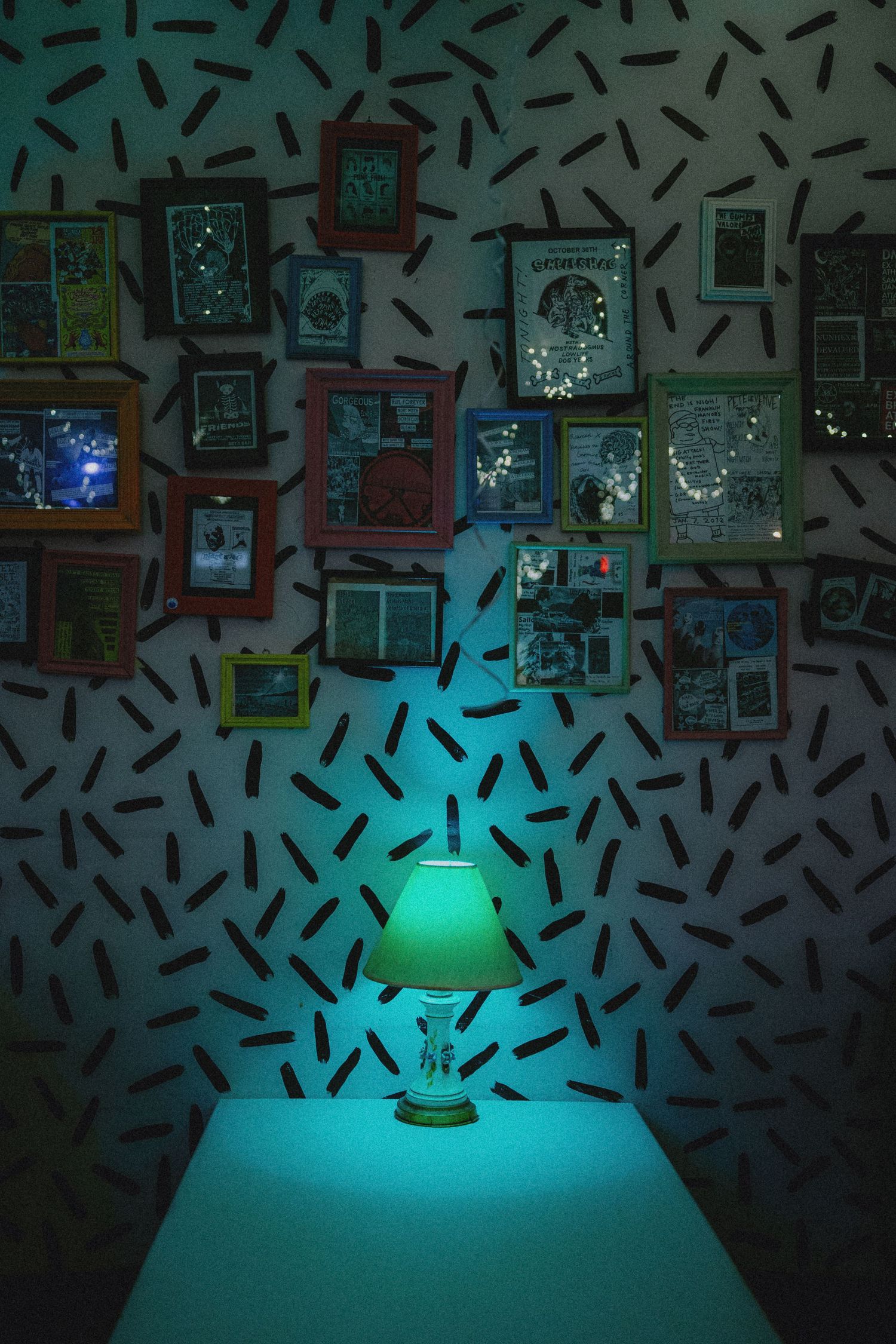 Glowing teal lamp on a table illuminates a gallery wall with colorful frames on a patterned wallpaper background.