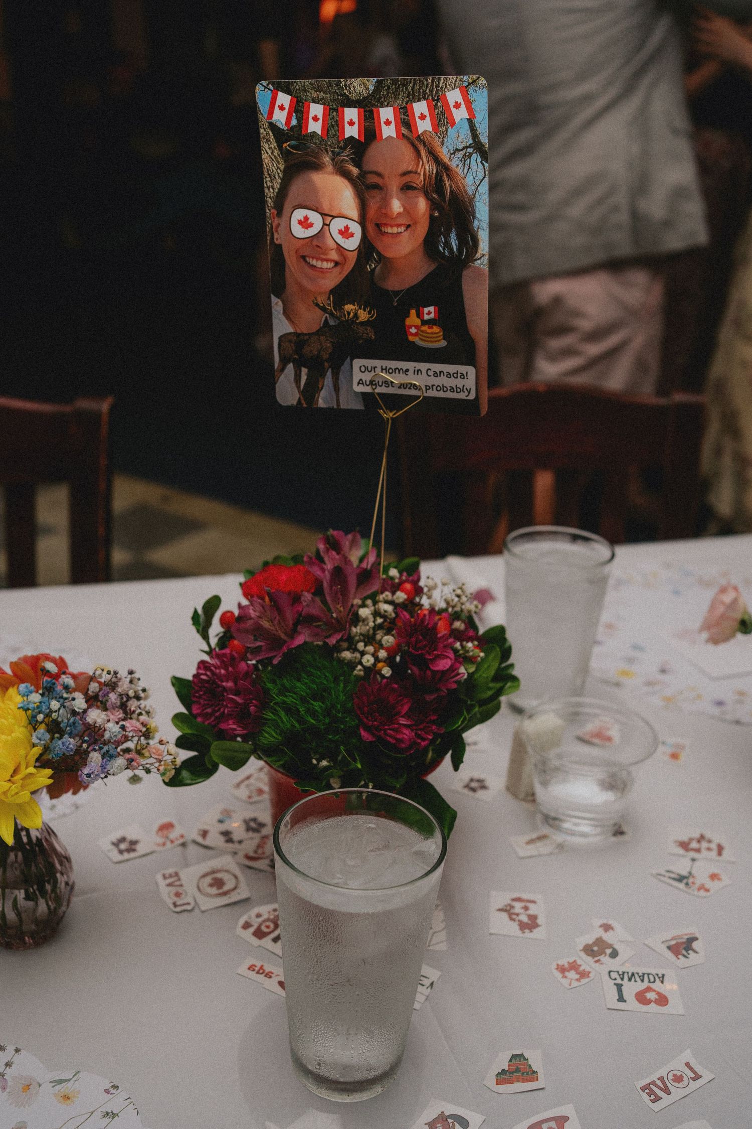 Wedding table centerpiece with colorful flowers and a photo of two women with Canadian flag glasses on a wire stand.