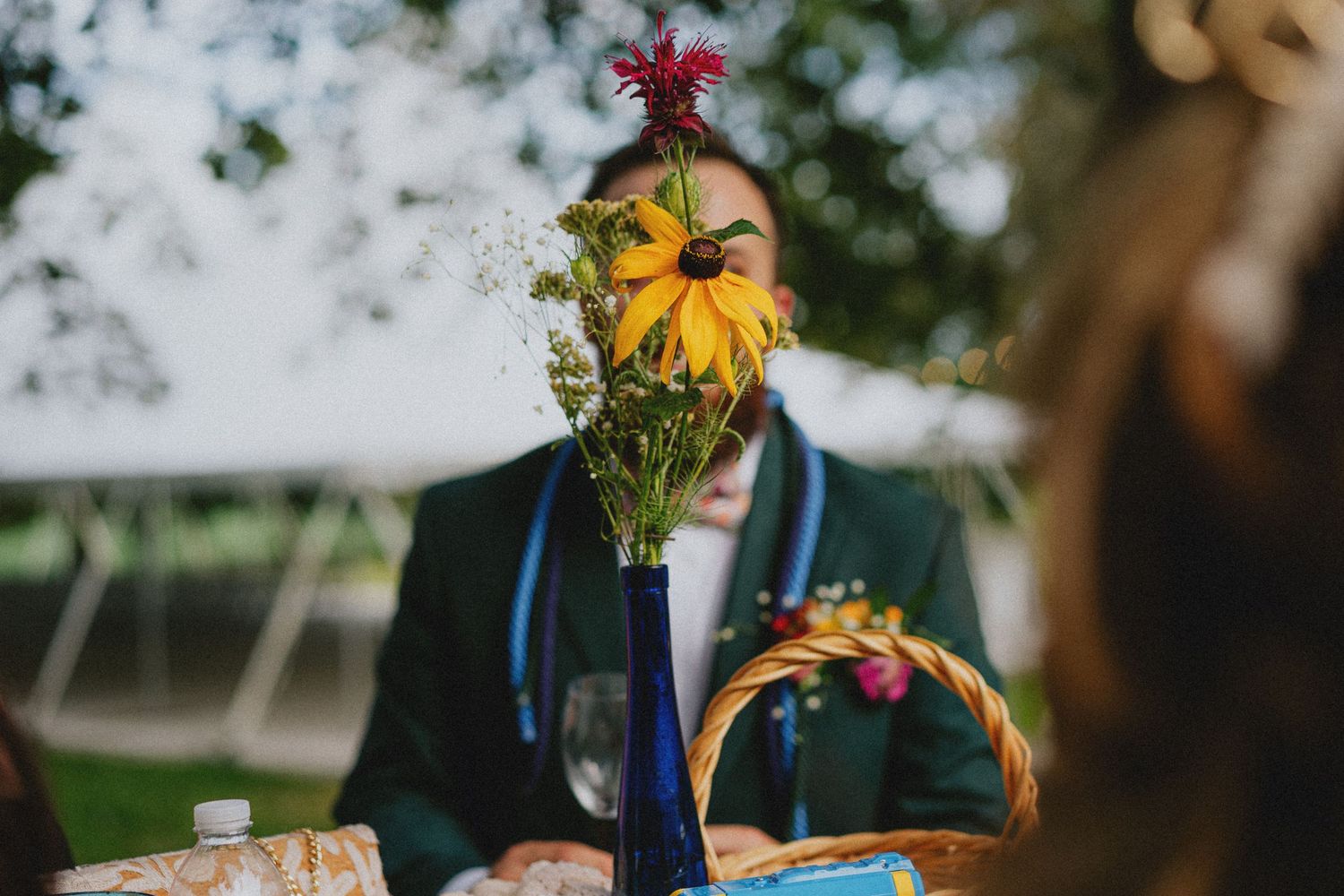 A vibrant floral centerpiece with yellow and red wildflowers in a blue vase at an outdoor wedding reception.