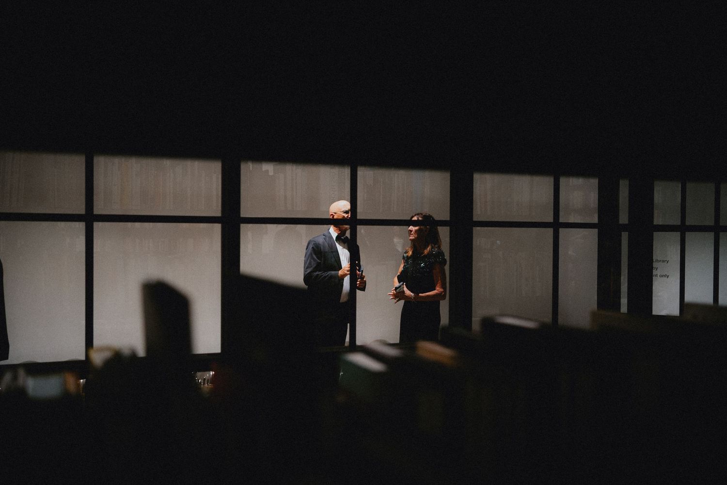 Two people stand in a dimly lit room with large windows, engaged in conversation in a dramatic, moody setting.
