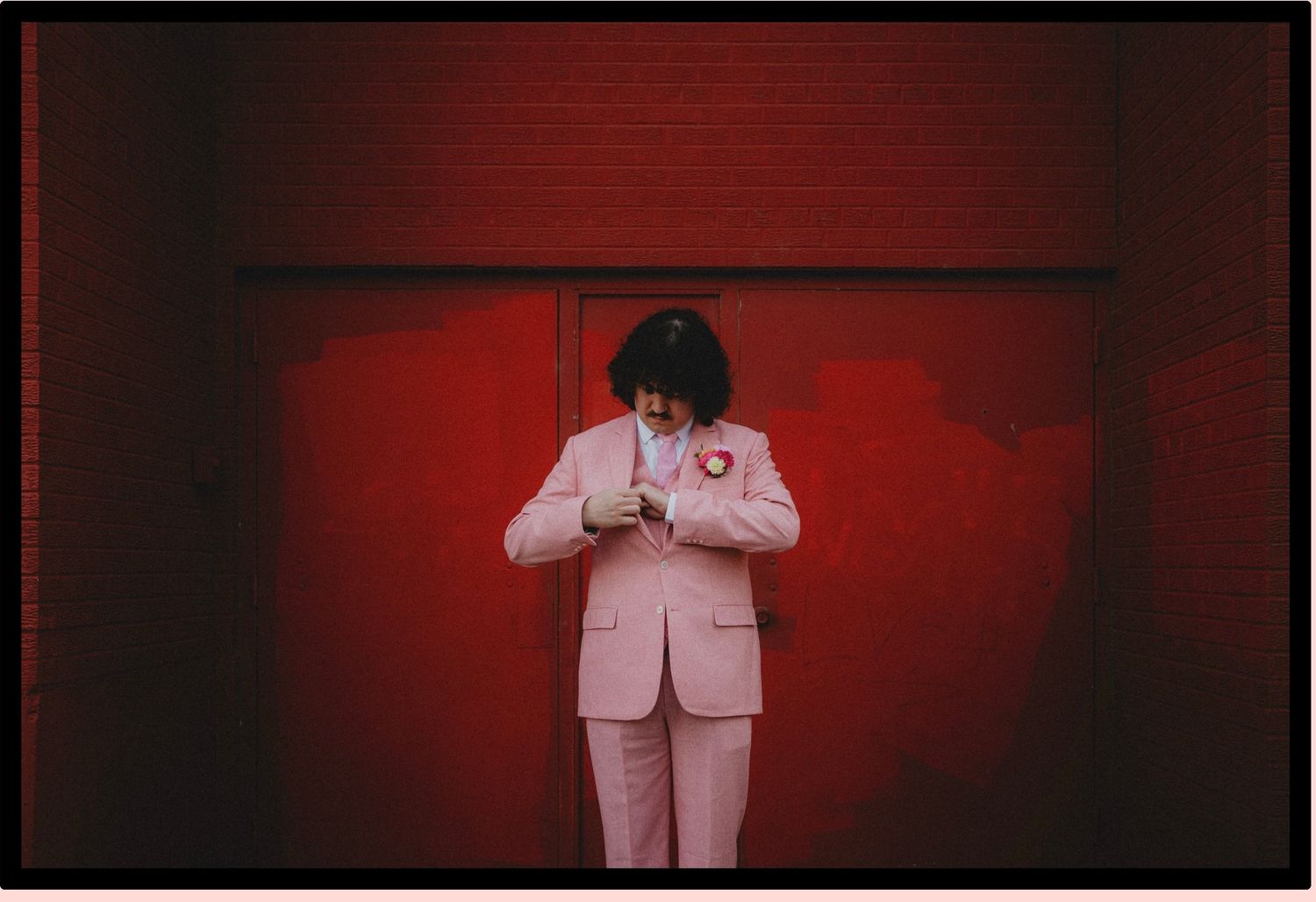 Person in pink suit standing against a red wall, looking down at a heart pin on their lapel.
