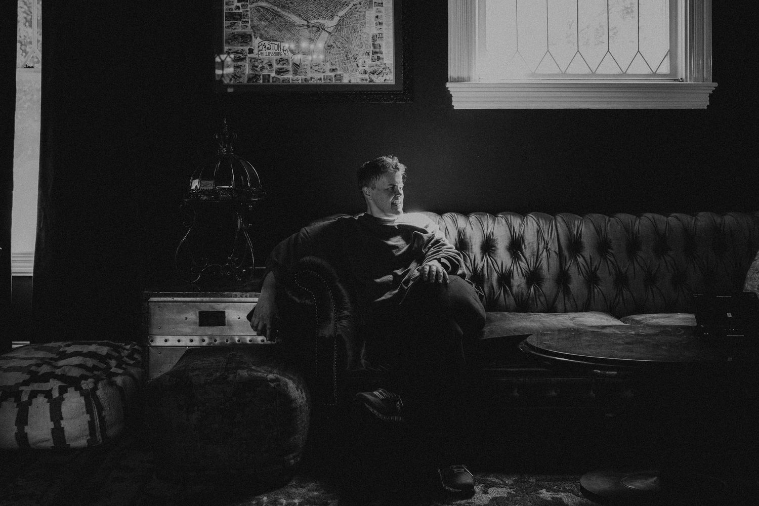 Black and white photo of a man sitting alone on a tufted sofa in a dimly lit, moody interior room.