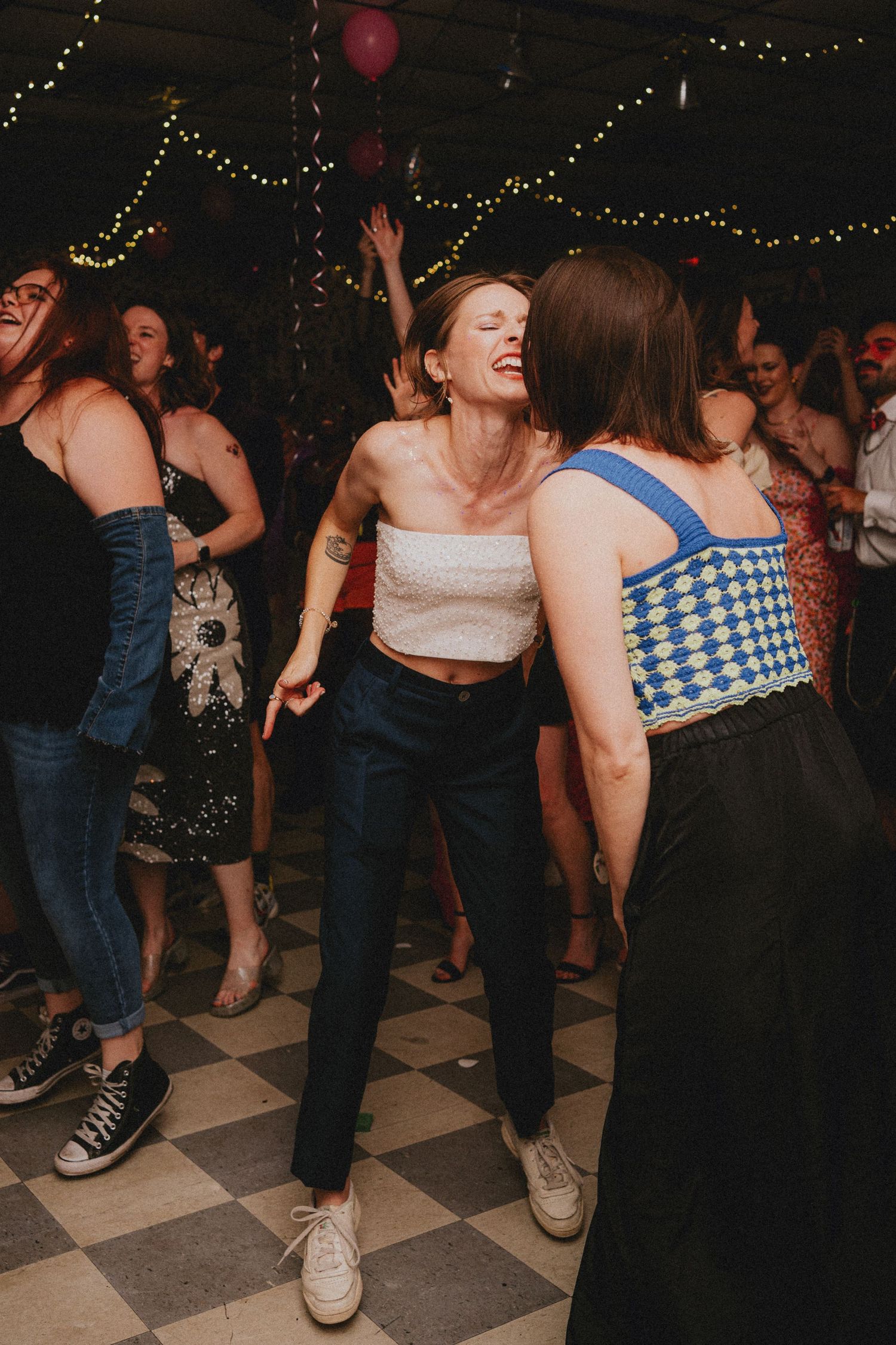 Two women dancing joyfully at a party on a checkered dance floor with string lights in the background.