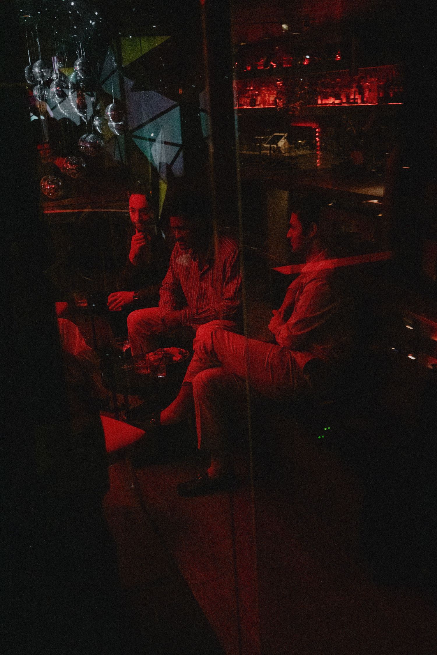 Three men relaxing in a dimly lit bar lounge with red ambient lighting and a stocked bar in the background.