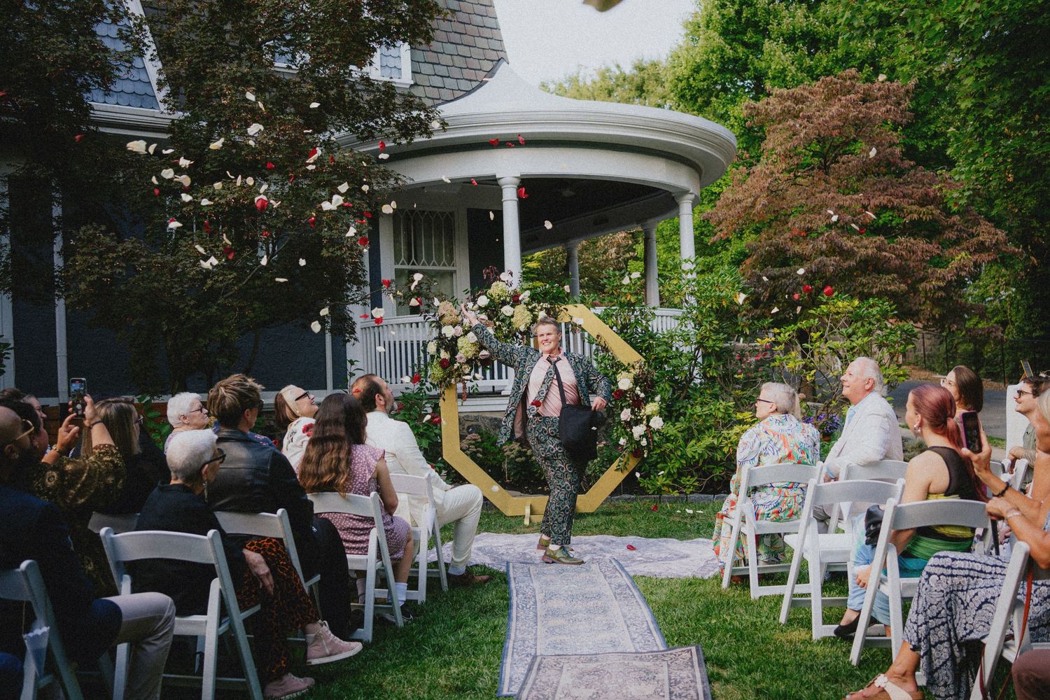 Outdoor wedding ceremony in a garden with guests seated in white chairs facing a hexagonal floral arch and a groom walking down the aisle.