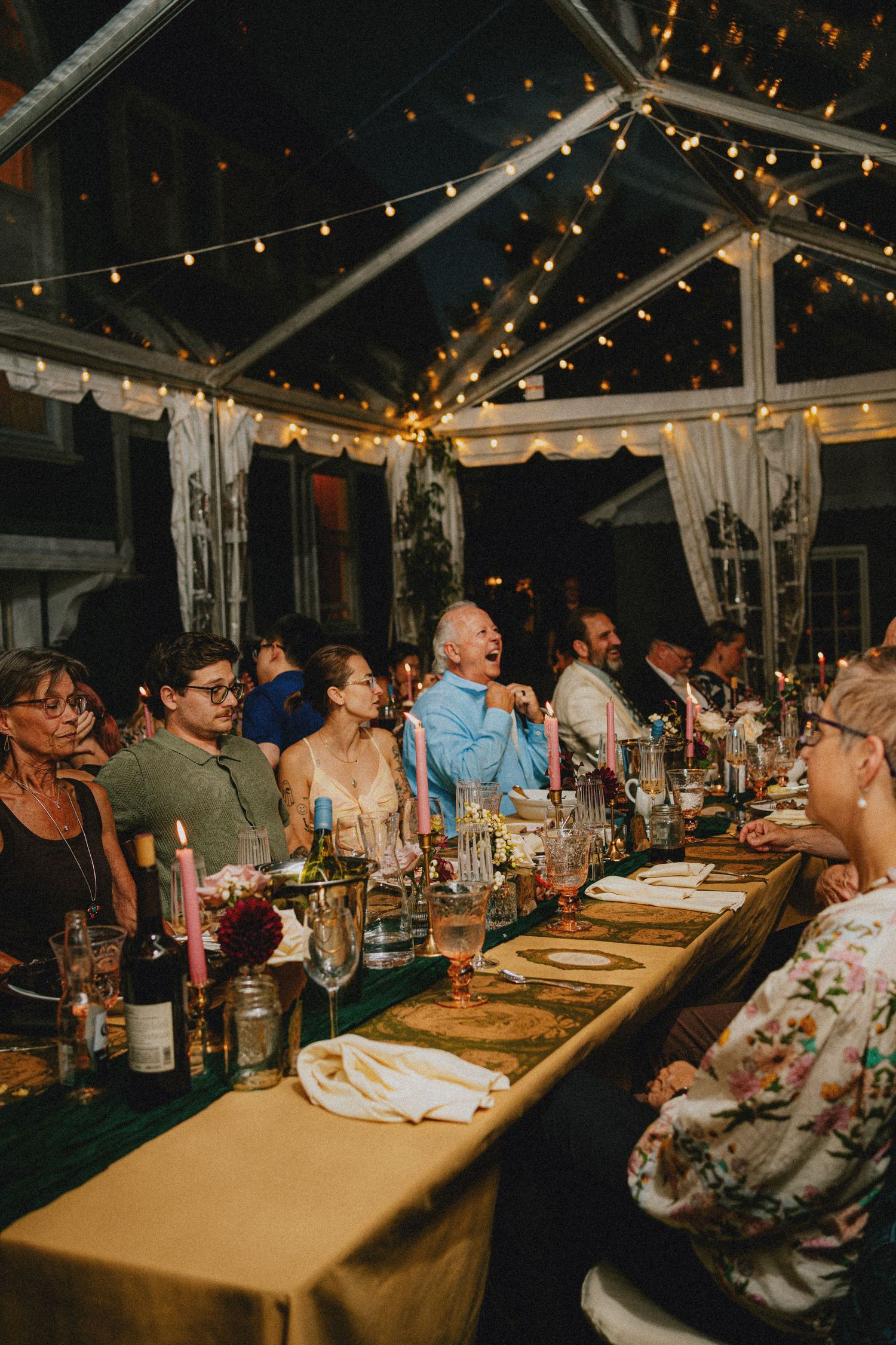 Guests laughing and dining under string lights at an elegant outdoor tented wedding reception at night.