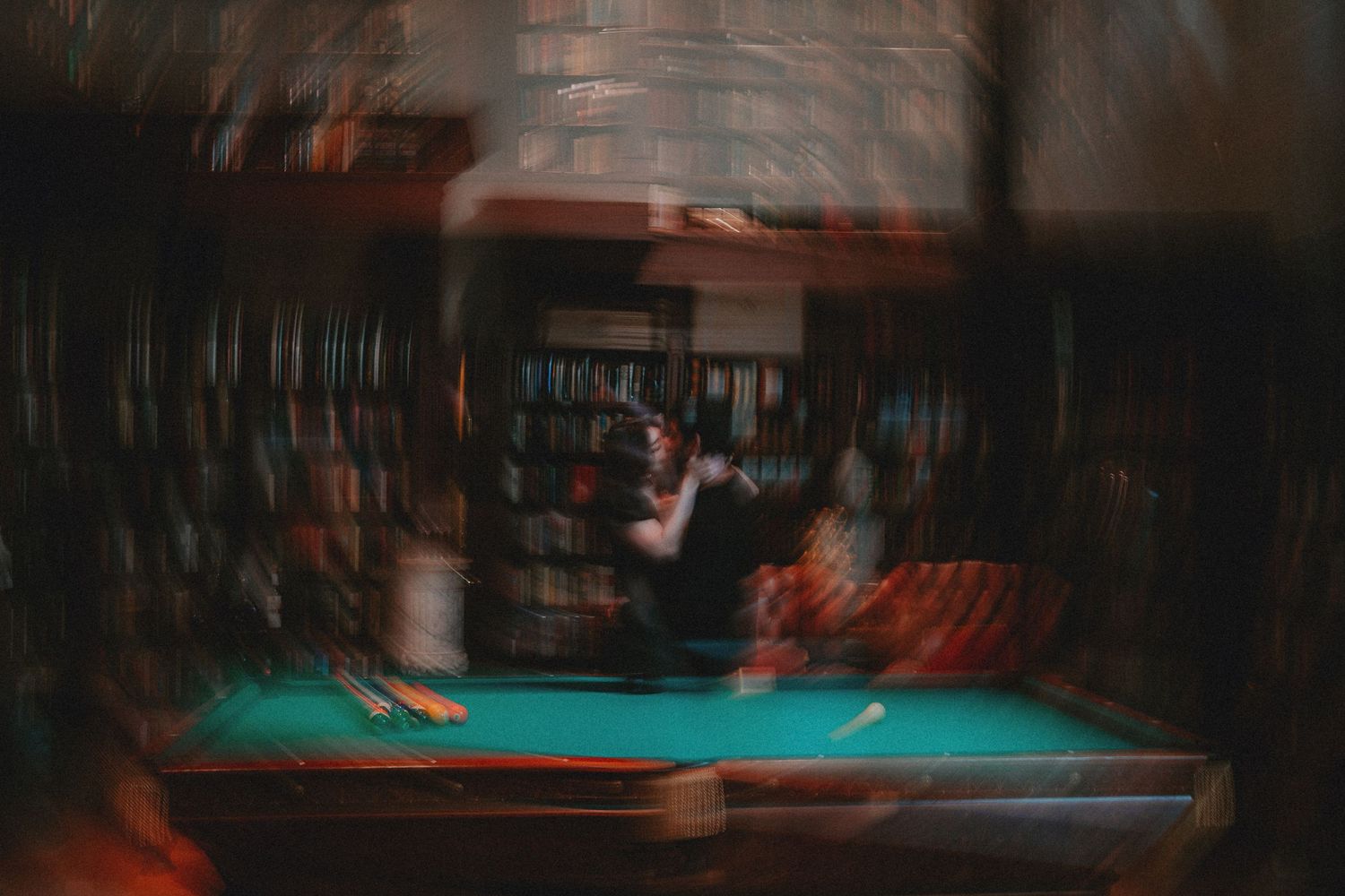 Blurry long-exposure photo of a person near a pool table in a dimly lit room with bookshelves in the background.