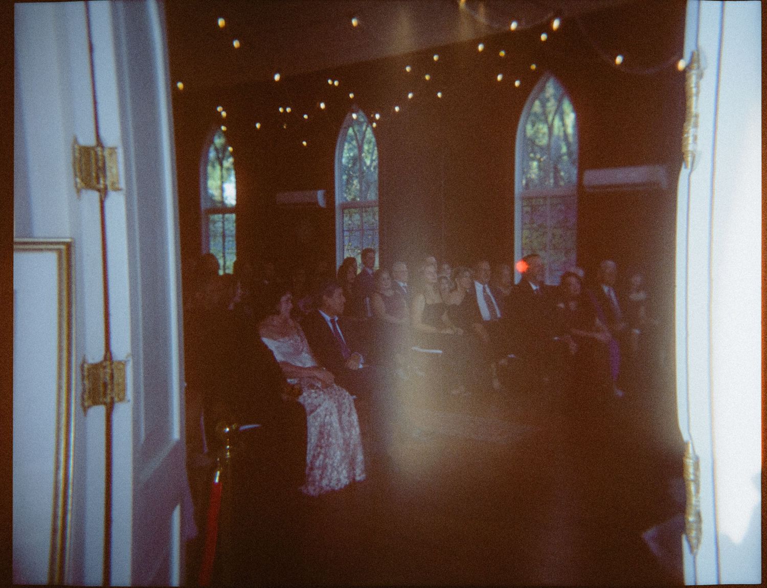 Guests seated in a dimly lit chapel with arched windows and string lights during an indoor wedding ceremony.