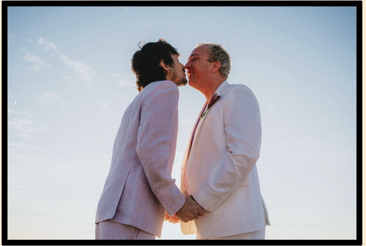 Two grooms in white suits share a kiss outdoors under a blue sky on their wedding day.