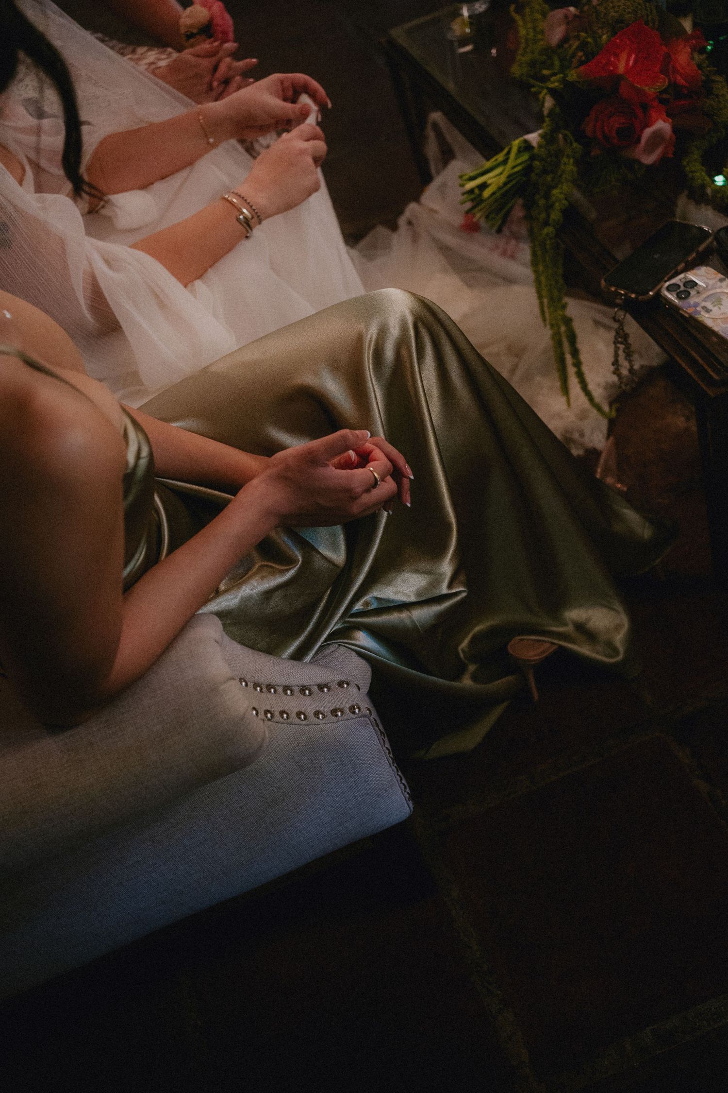 Two women in elegant satin and white gowns seated together, with a floral arrangement nearby in a dimly lit setting.