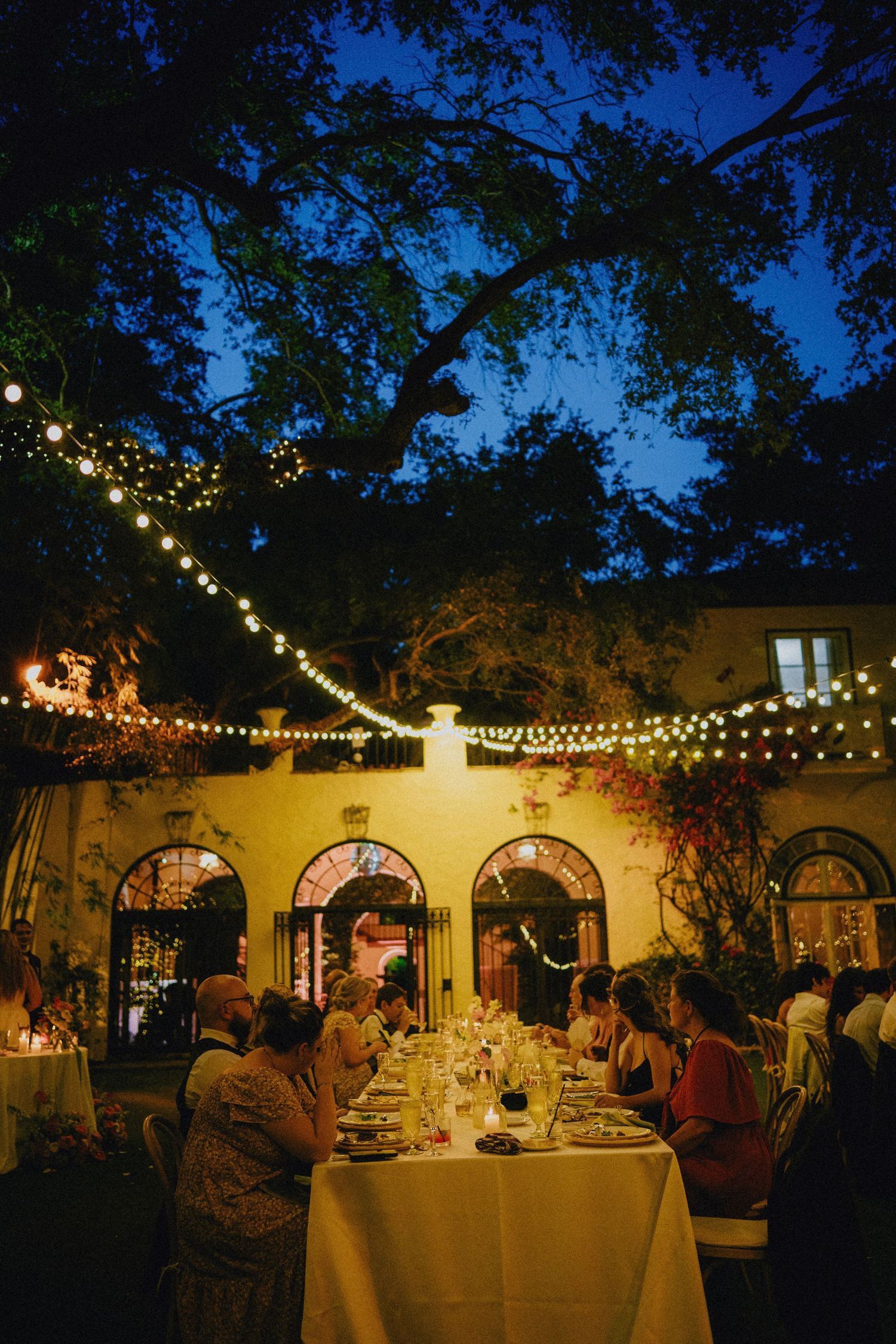 Outdoor evening wedding reception with string lights, long dining table, and guests under a large tree at a yellow villa.