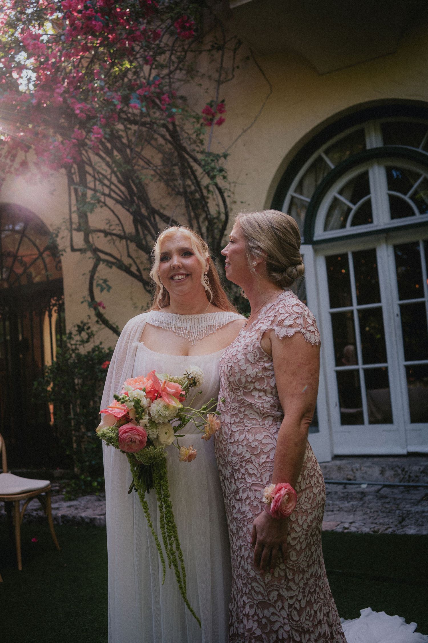 Bride in white cape gown holds pink bouquet beside older woman in lace dress outside elegant venue.