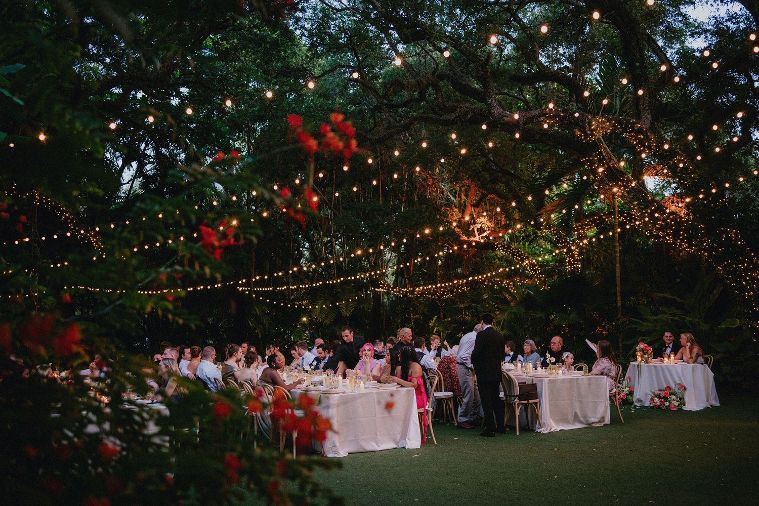 Outdoor wedding reception under string lights and red lanterns in a lush garden at dusk.