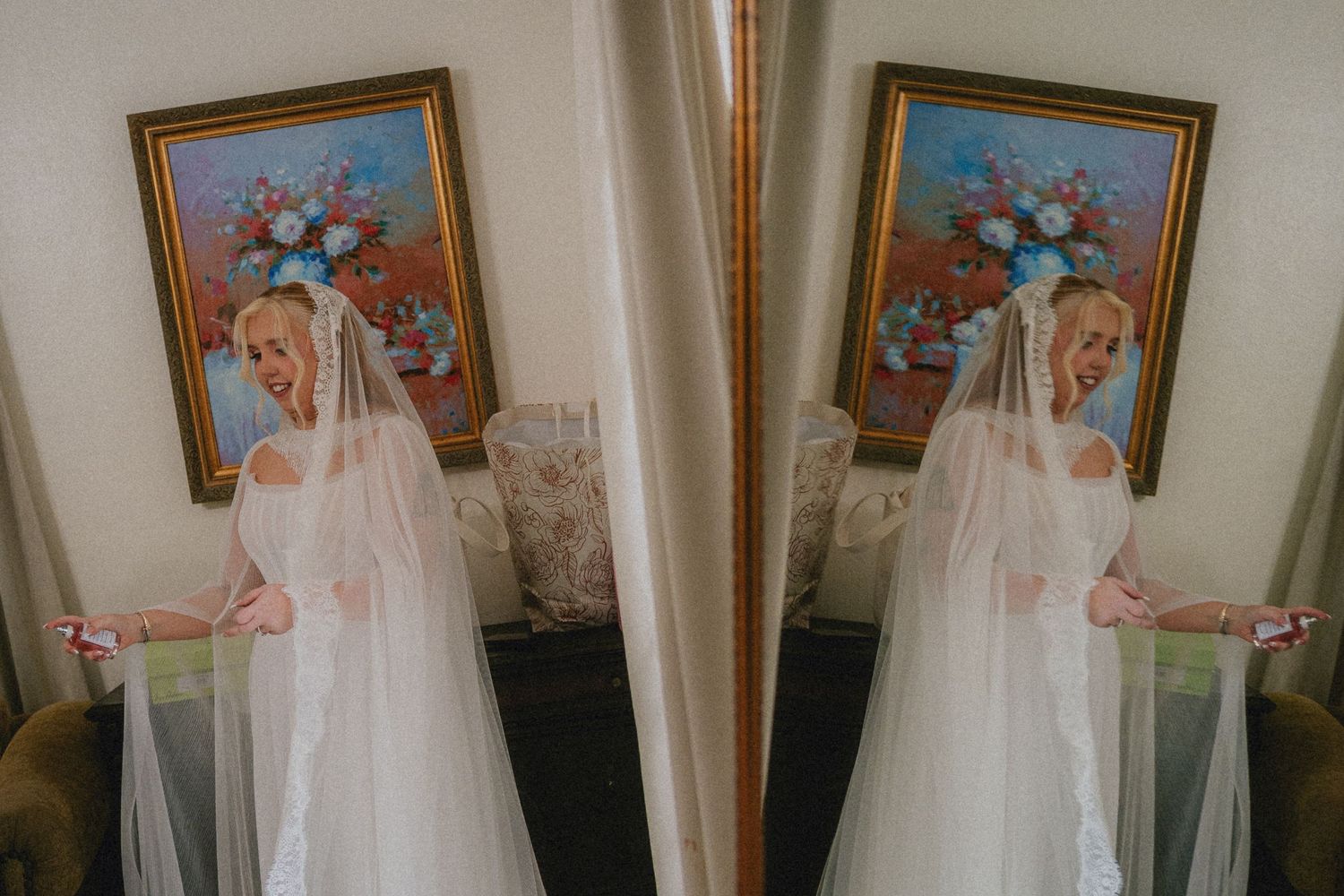 Bride in white wedding dress and veil poses near curtains with floral painting, reflected in mirror.