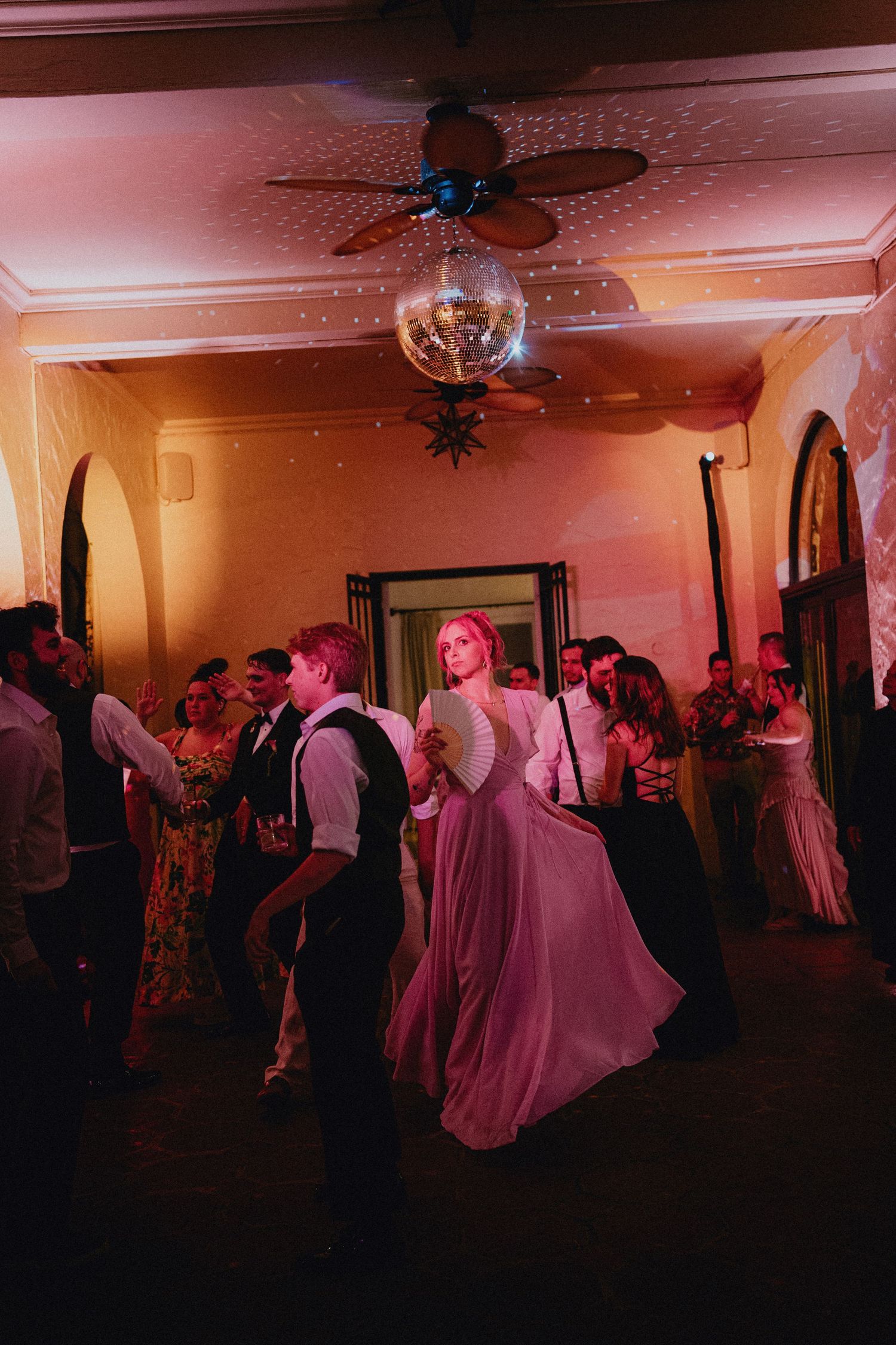 Bride in pink gown dances with groom under disco ball at elegant wedding reception with warm lighting.