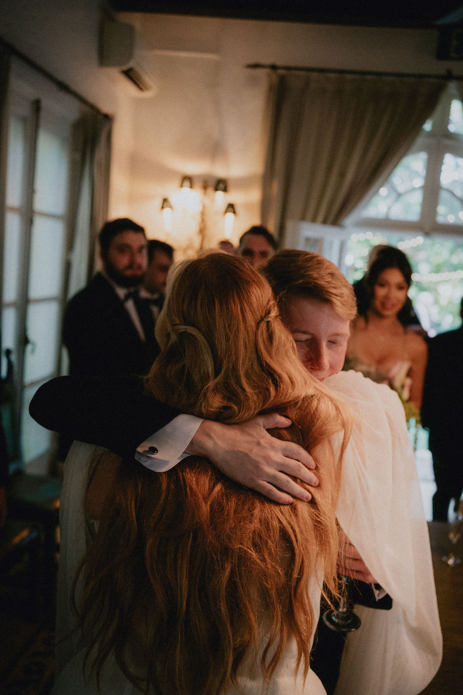 A bride and groom share a warm embrace at their wedding reception, surrounded by guests in elegant attire.
