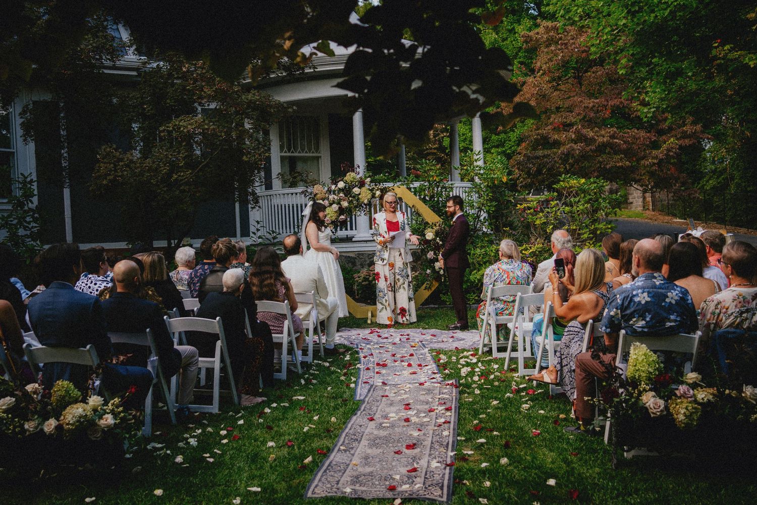Outdoor wedding ceremony on a lush green lawn with guests seated in white chairs, floral arch, and rose petal aisle.