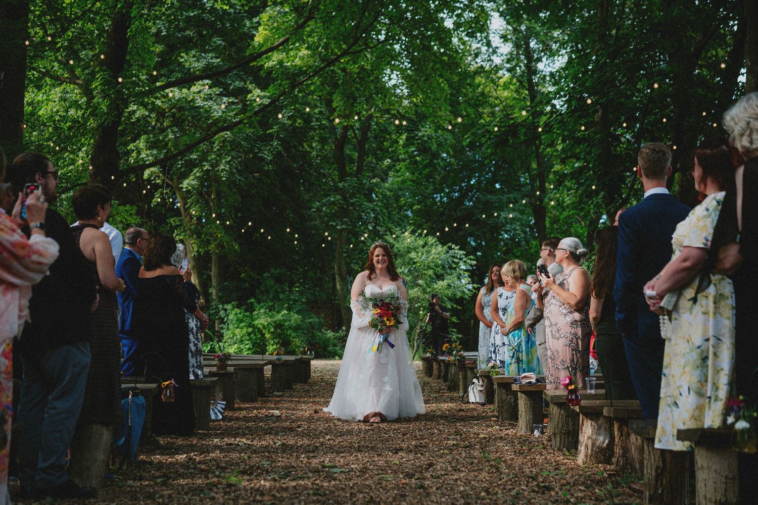 Bride walks down outdoor woodland aisle lined with guests and string lights at a romantic forest wedding ceremony.