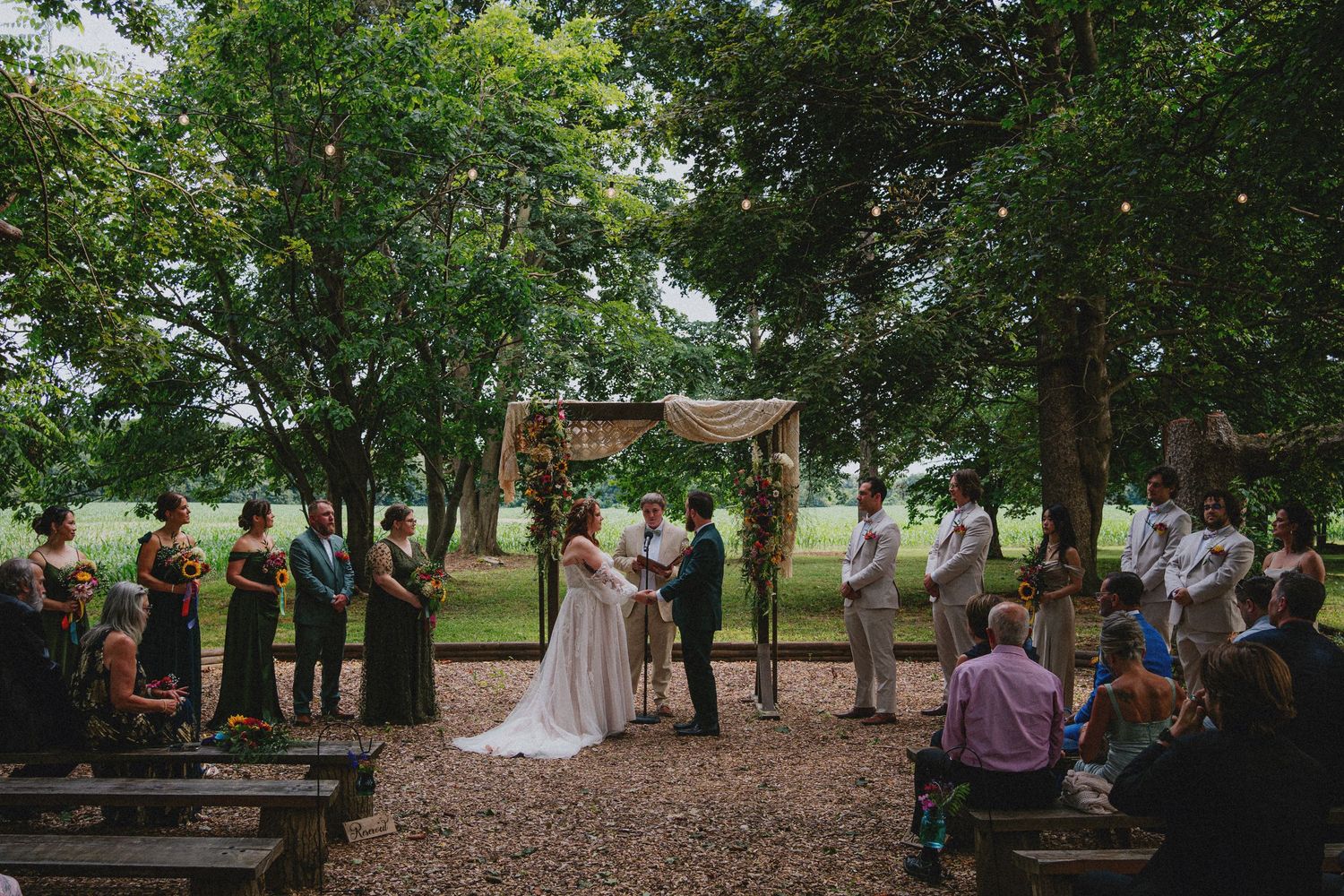 Outdoor wedding ceremony under trees with couple at rustic wooden arch, guests seated on benches in lush green setting.