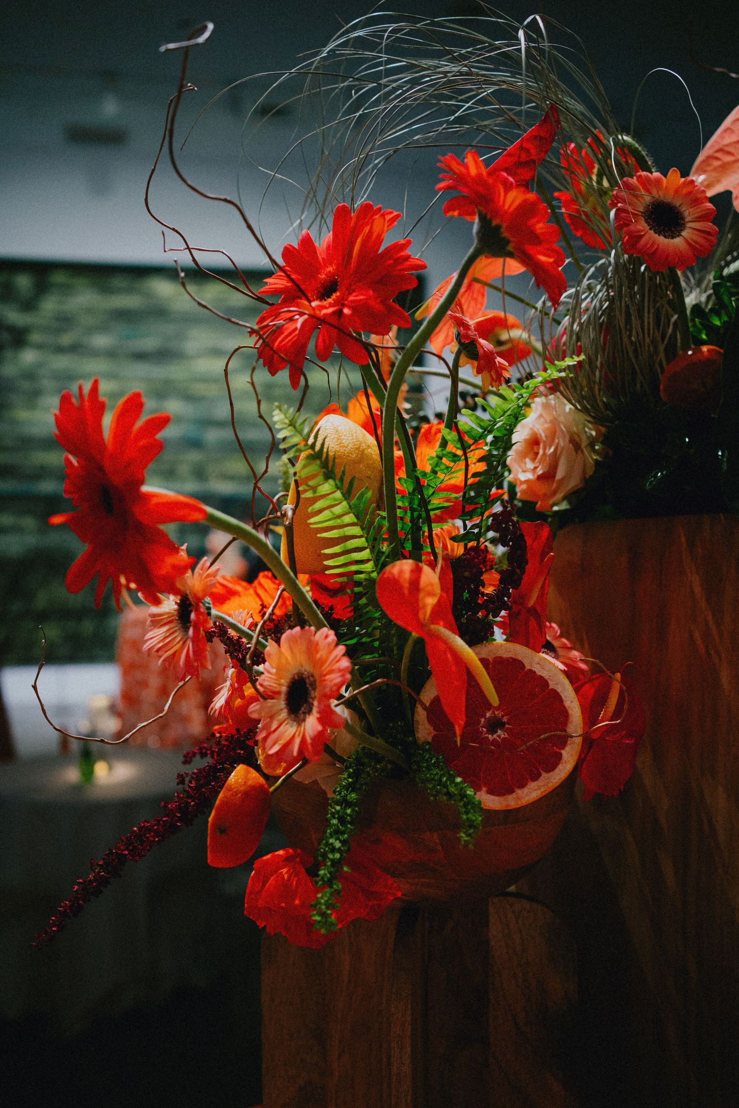 Vibrant red gerbera daisy floral arrangement with anthurium, grapefruit slices, and greenery on a wooden stand.