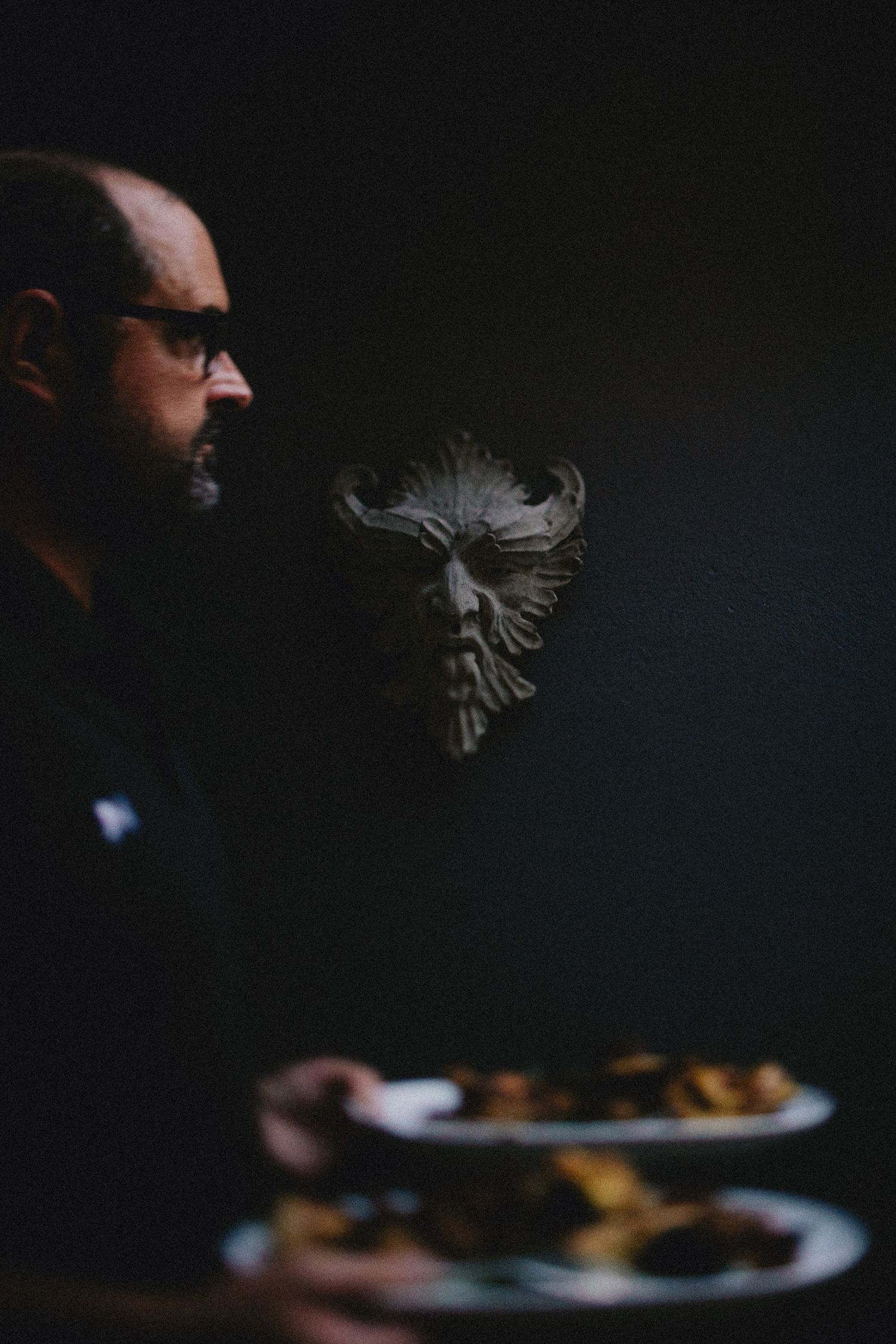 Man with glasses beside a decorative demon face sculpture in a dark, moody setting with a plate of food nearby.