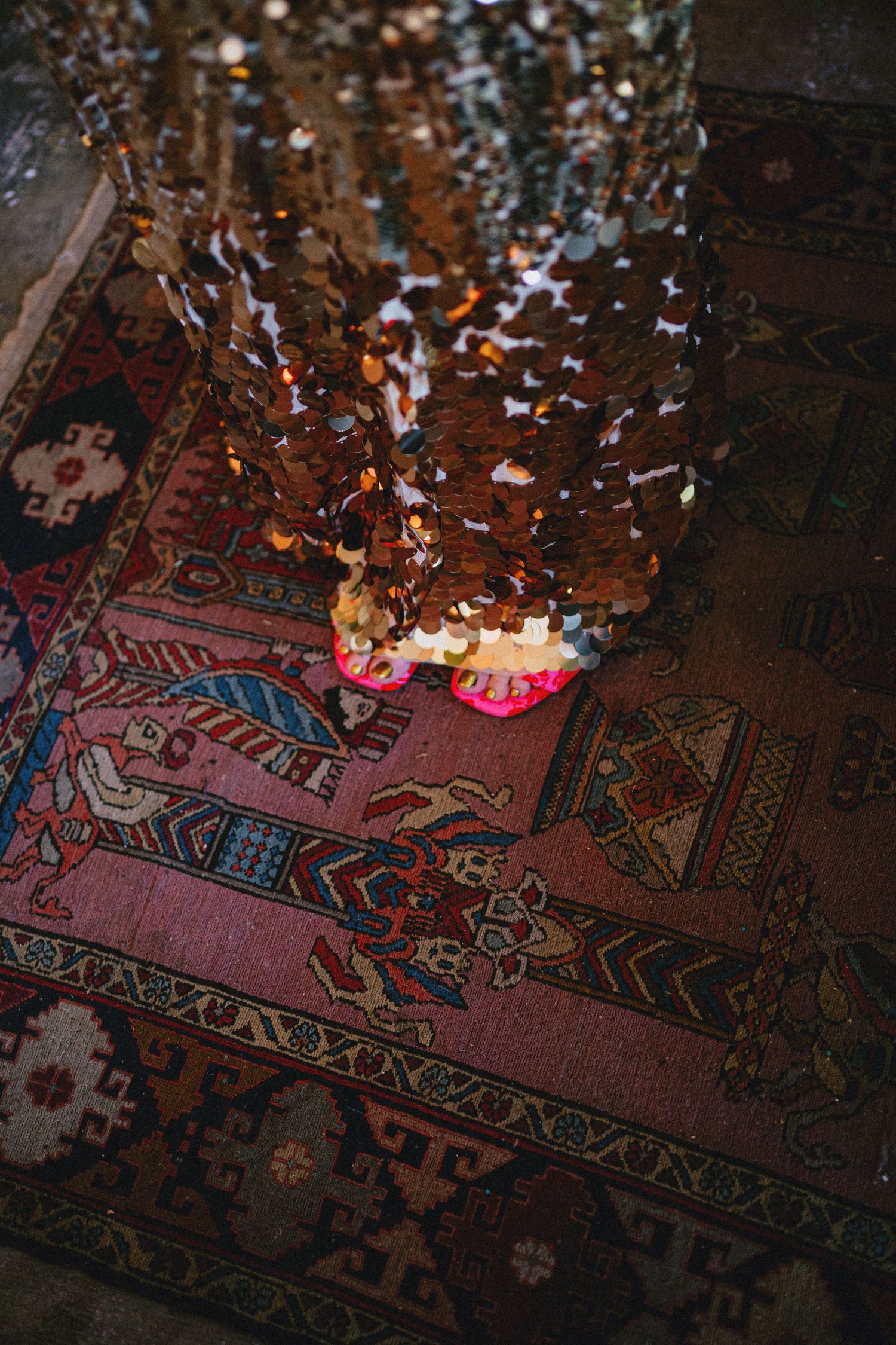 Sequined dress hem with pink shoes on ornate Persian rug, sparkling gold sequins catching light.
