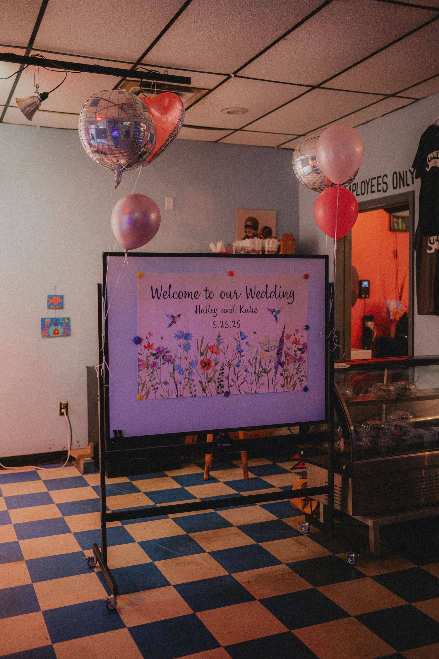 Wedding welcome sign on TV display with pink balloons and disco ball decorations on checkered floor.