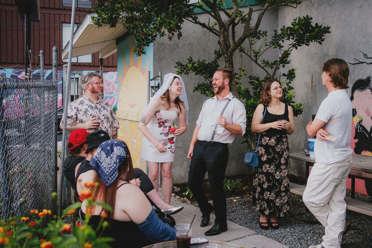 Group of friends socializing outdoors at a casual garden party, standing and sitting near a colorful mural wall.