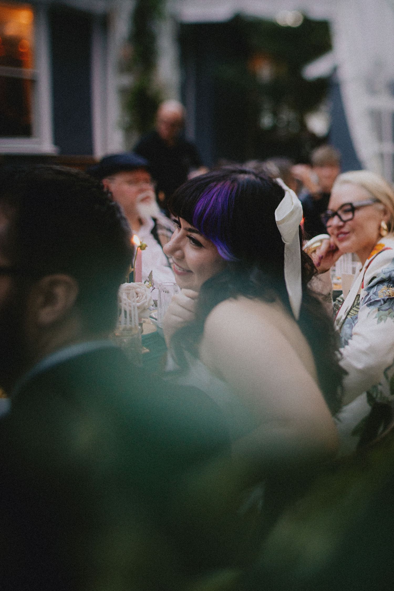 A woman with purple hair and white ribbon smiles at an outdoor evening wedding reception gathering.