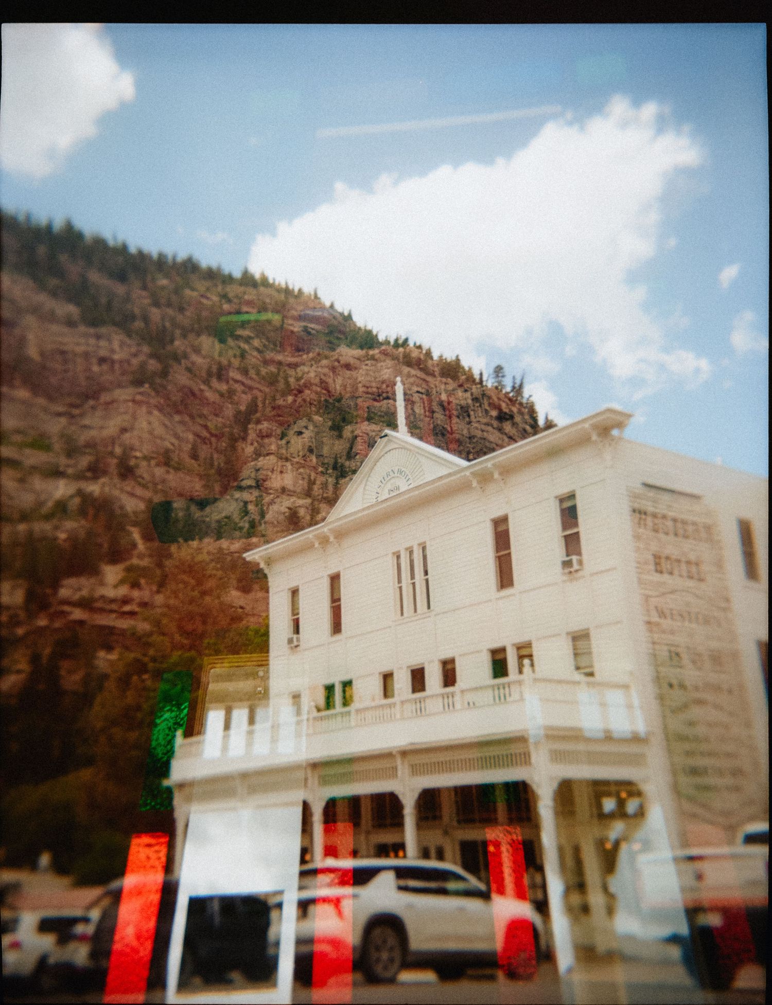 Double-exposed film photo of a white Victorian building against red rock cliffs and blue sky in a mountain town.