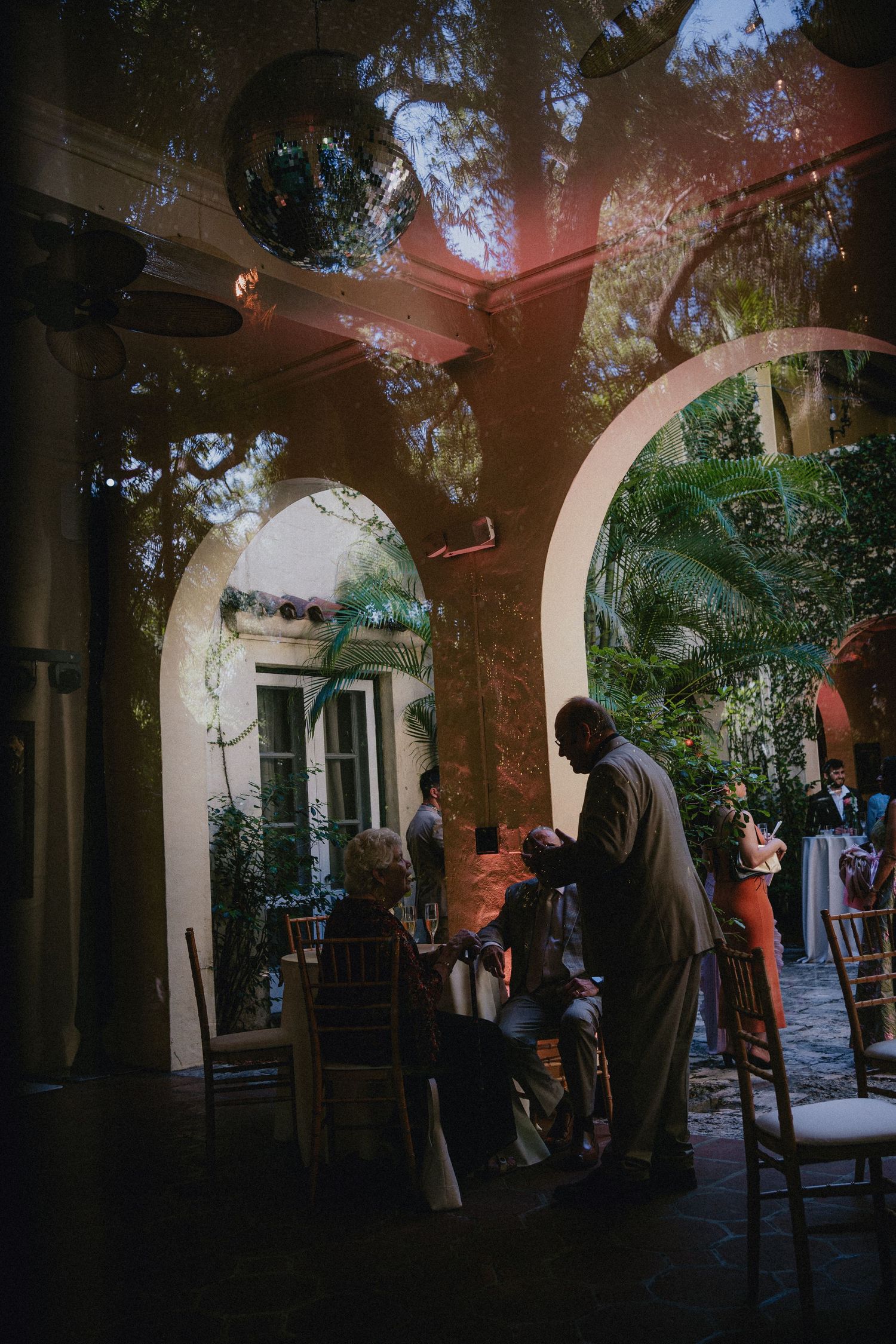 Elegant outdoor courtyard event with arched doorways, lush trees, and guests gathered under warm ambient lighting.