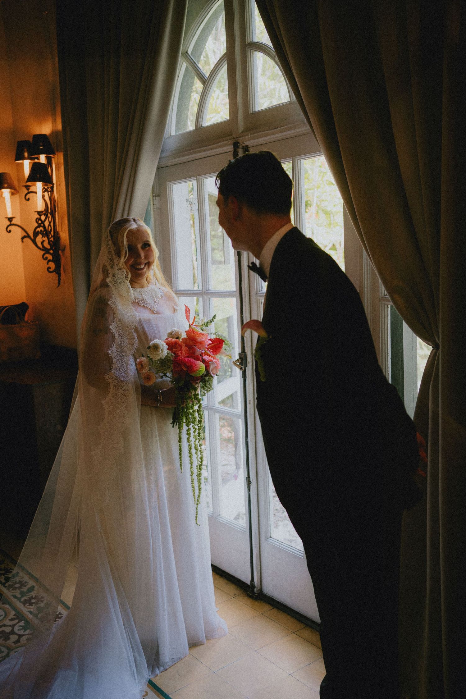Bride in lace gown holding colorful bouquet smiles at groom in black tuxedo by arched window with draping curtains.