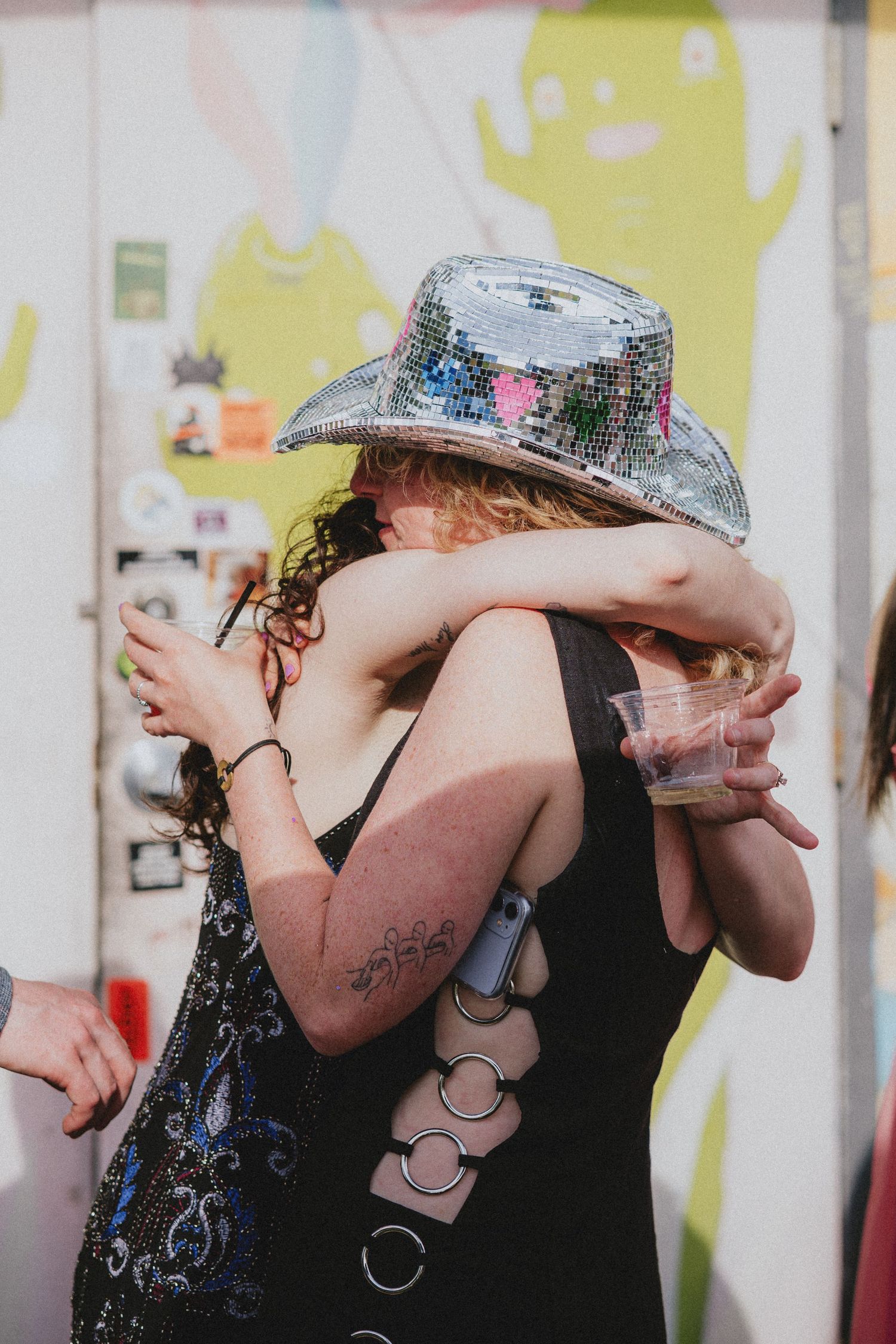 Two friends hugging at a festival, one wearing a silver disco ball cowboy hat and holding a drink.