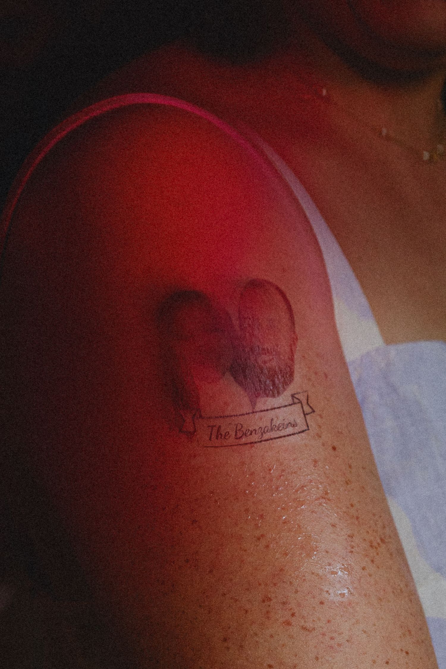 A shoulder tattoo of two fingerprints forming a heart shape with 'The Benjakeens' banner beneath, photographed in red light.