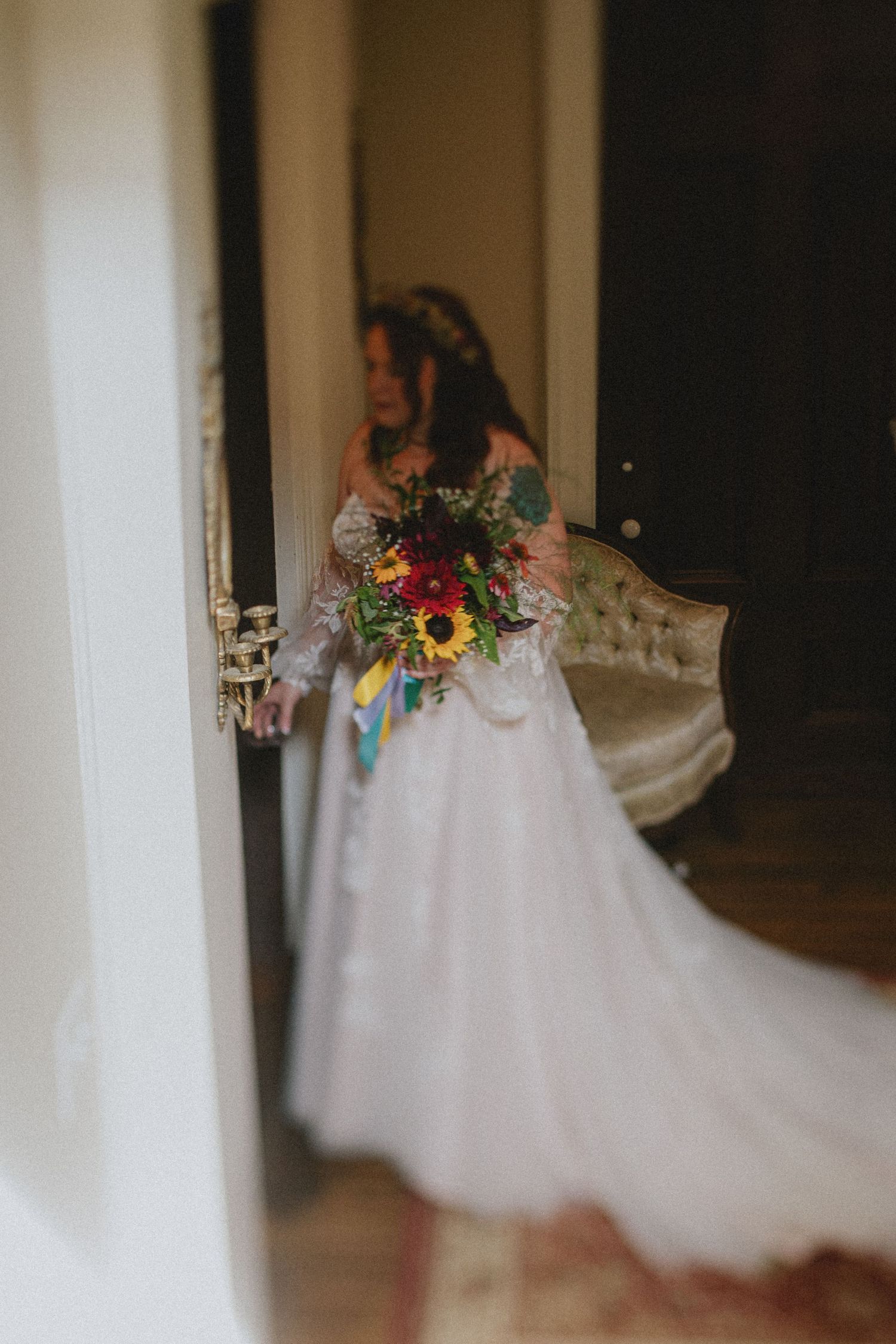 Bride in white gown holding colorful sunflower bouquet standing in doorway of elegant room.