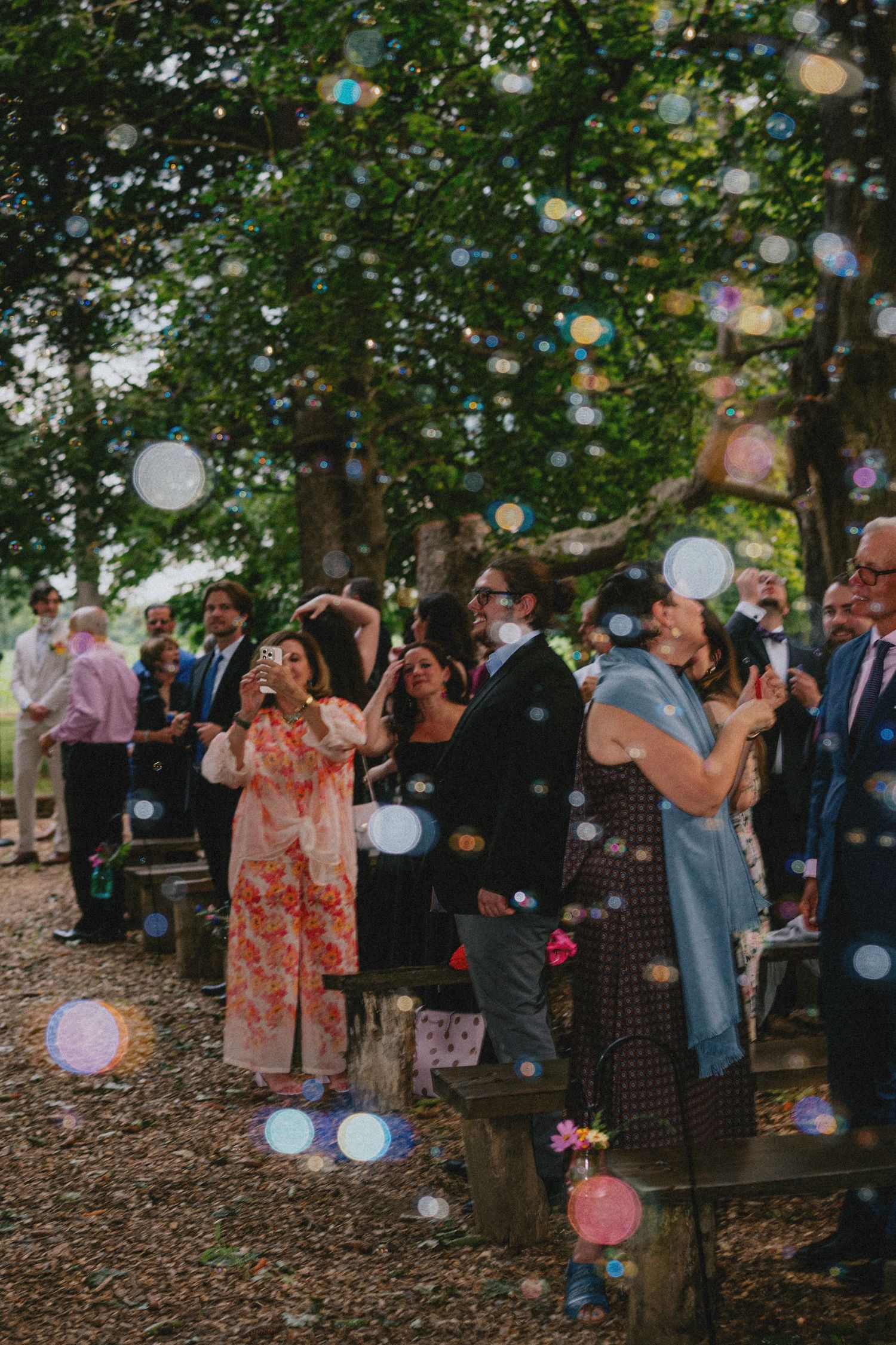 Guests blowing bubbles at an outdoor wedding ceremony surrounded by lush green trees.