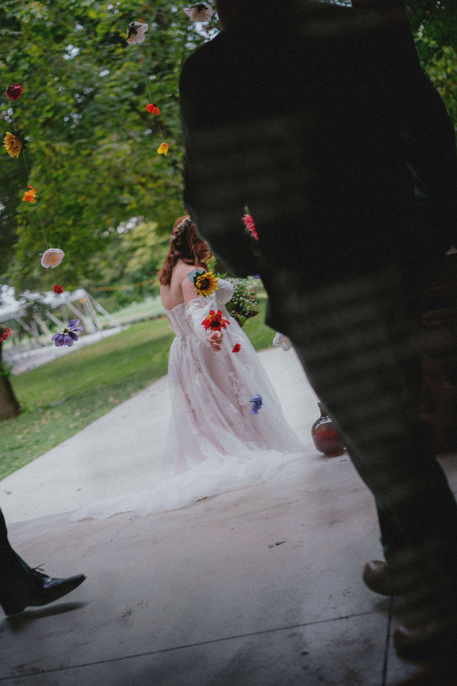 Bride in floral wedding gown holding sunflower bouquet walks through outdoor garden ceremony venue.