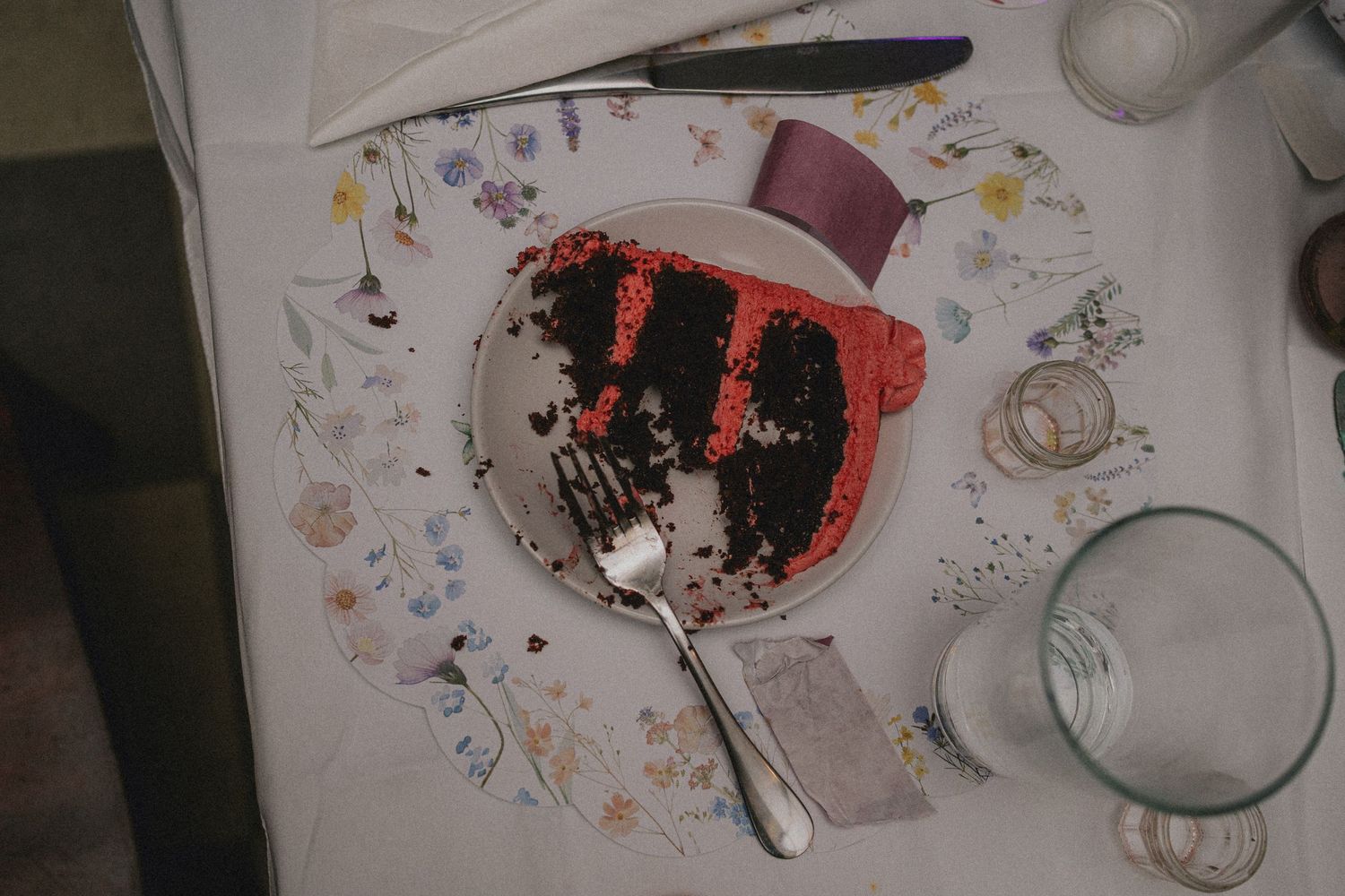 Partially eaten red velvet cake slice on a floral plate with a fork, surrounded by glasses on a white tablecloth.