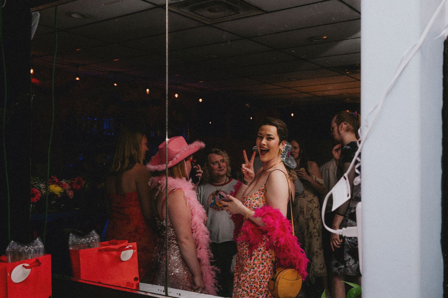 Women in colorful party outfits, including pink hat and feather boa, posing and flashing peace sign at festive event.