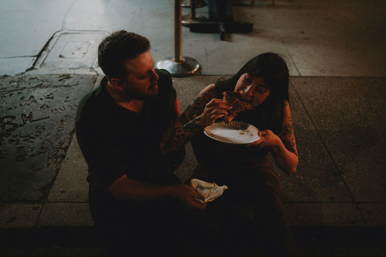 A tattooed couple sharing a meal together outdoors at night, sitting on steps in casual black clothing.