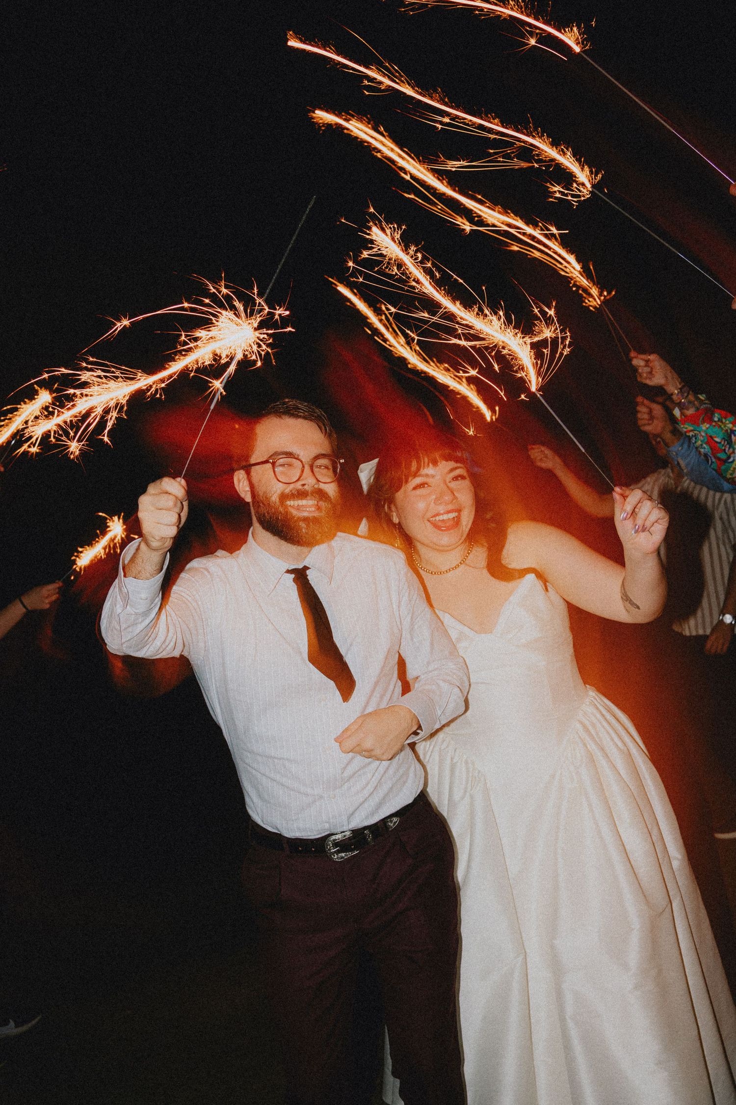 Joyful newlyweds walk through a sparkler tunnel at night, bride in white gown and groom in white shirt with dark tie.