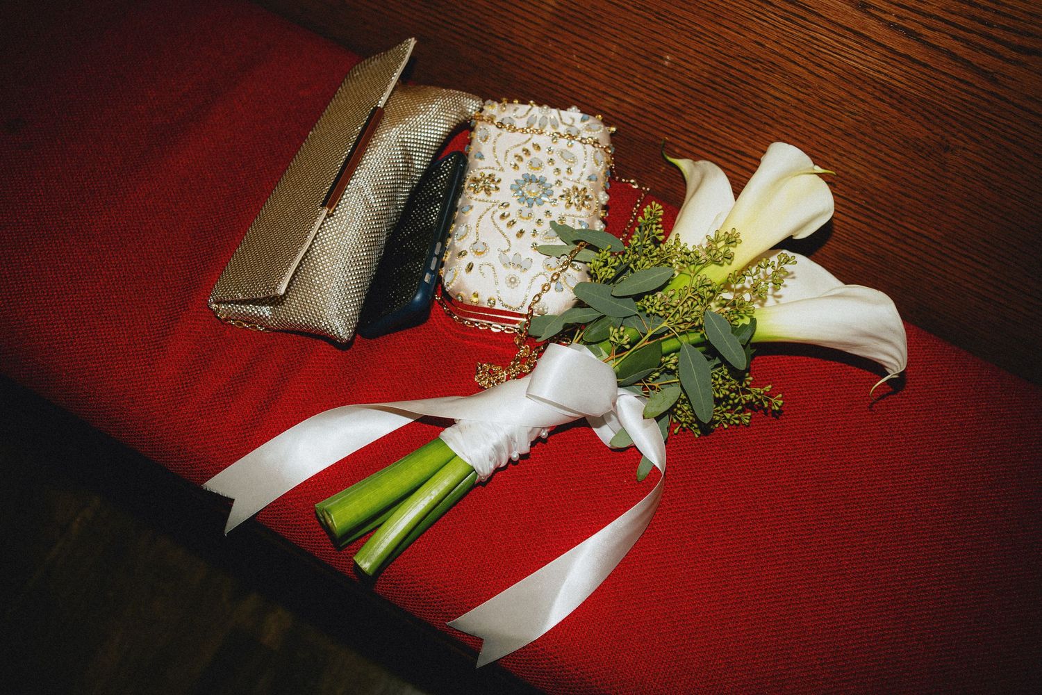 White calla lily wedding bouquet with ribbon on red velvet surface beside beaded clutch purse.