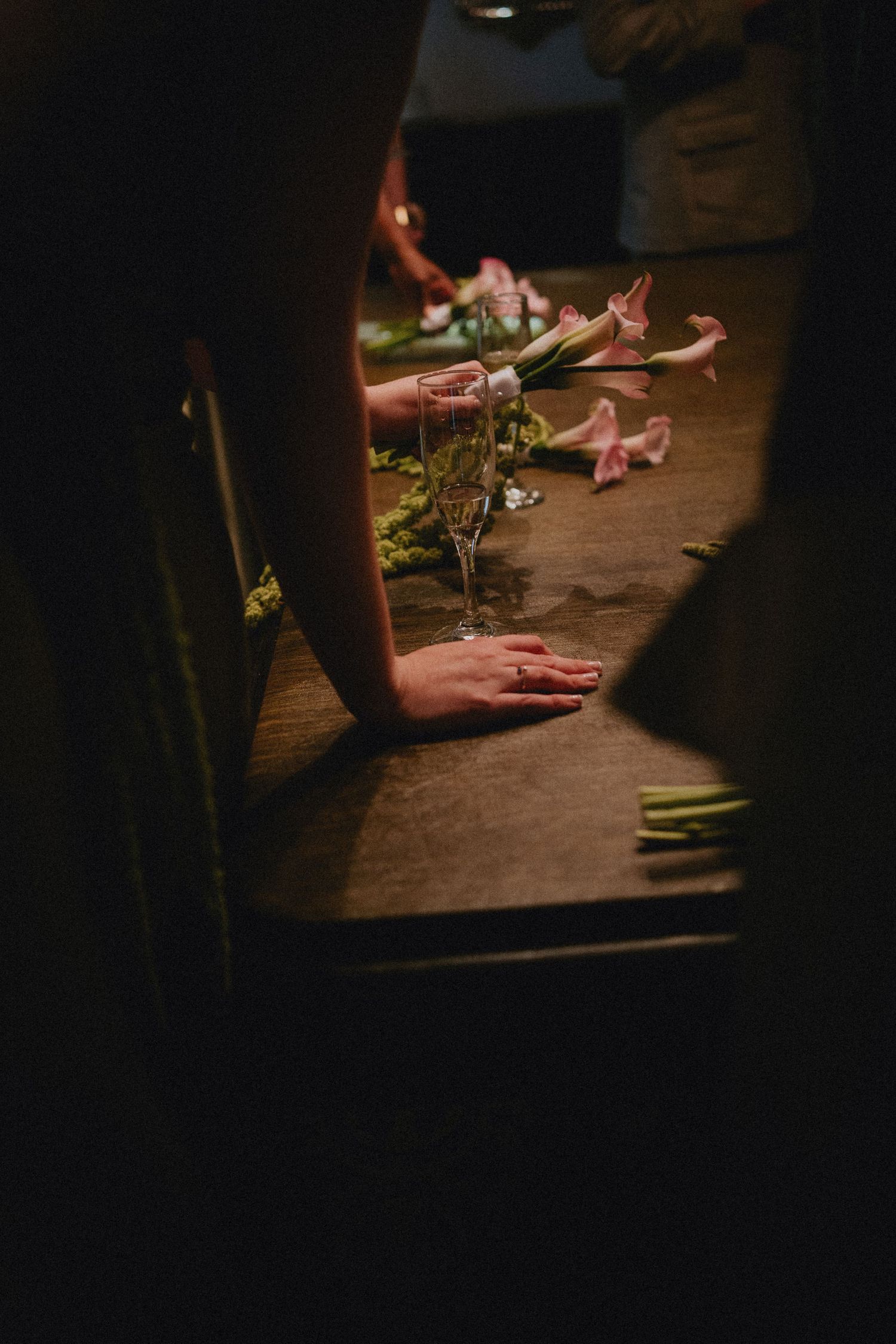 Hands resting on a wooden table with pink calla lilies and a glass vase in moody, dim lighting.