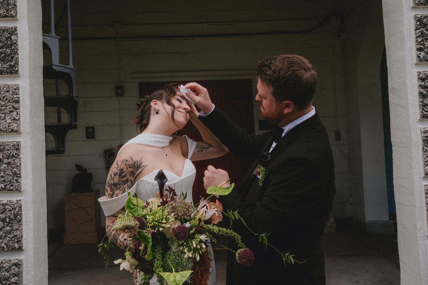 Groom adjusting bride's hair before wedding ceremony, bride holding lush floral bouquet outdoors.