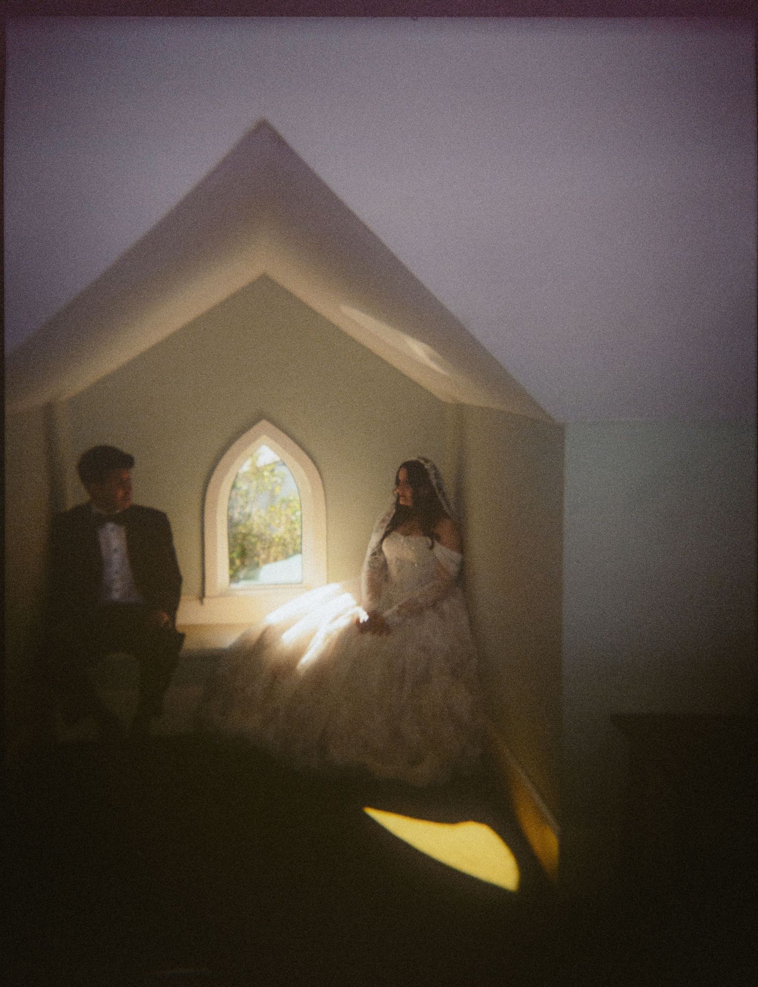 Bride and groom sitting inside a chapel alcove with gothic arch window, dramatic light casting warm glow on wedding dress.