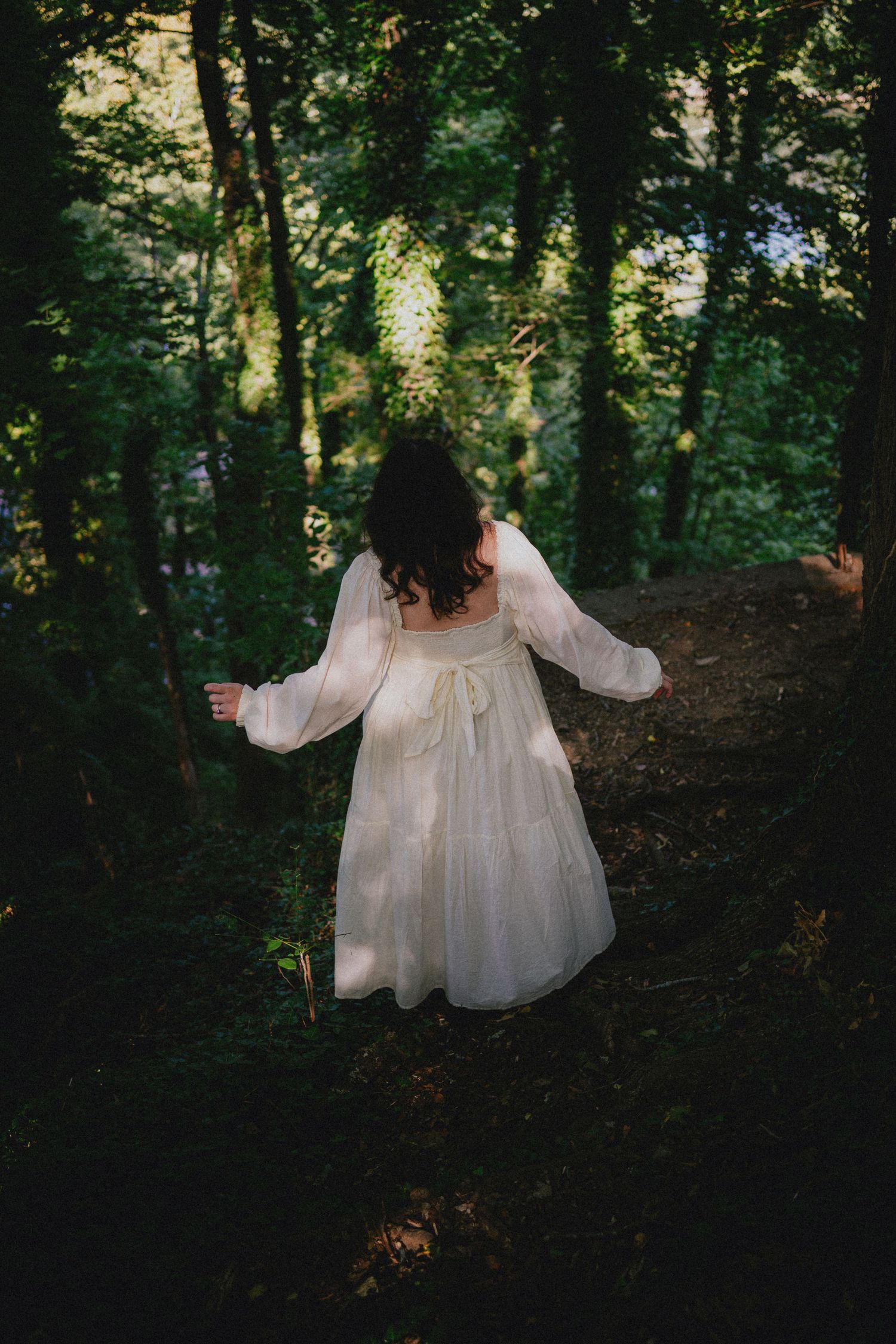 A woman in a flowing white dress walks through a lush green forest on a dirt path.