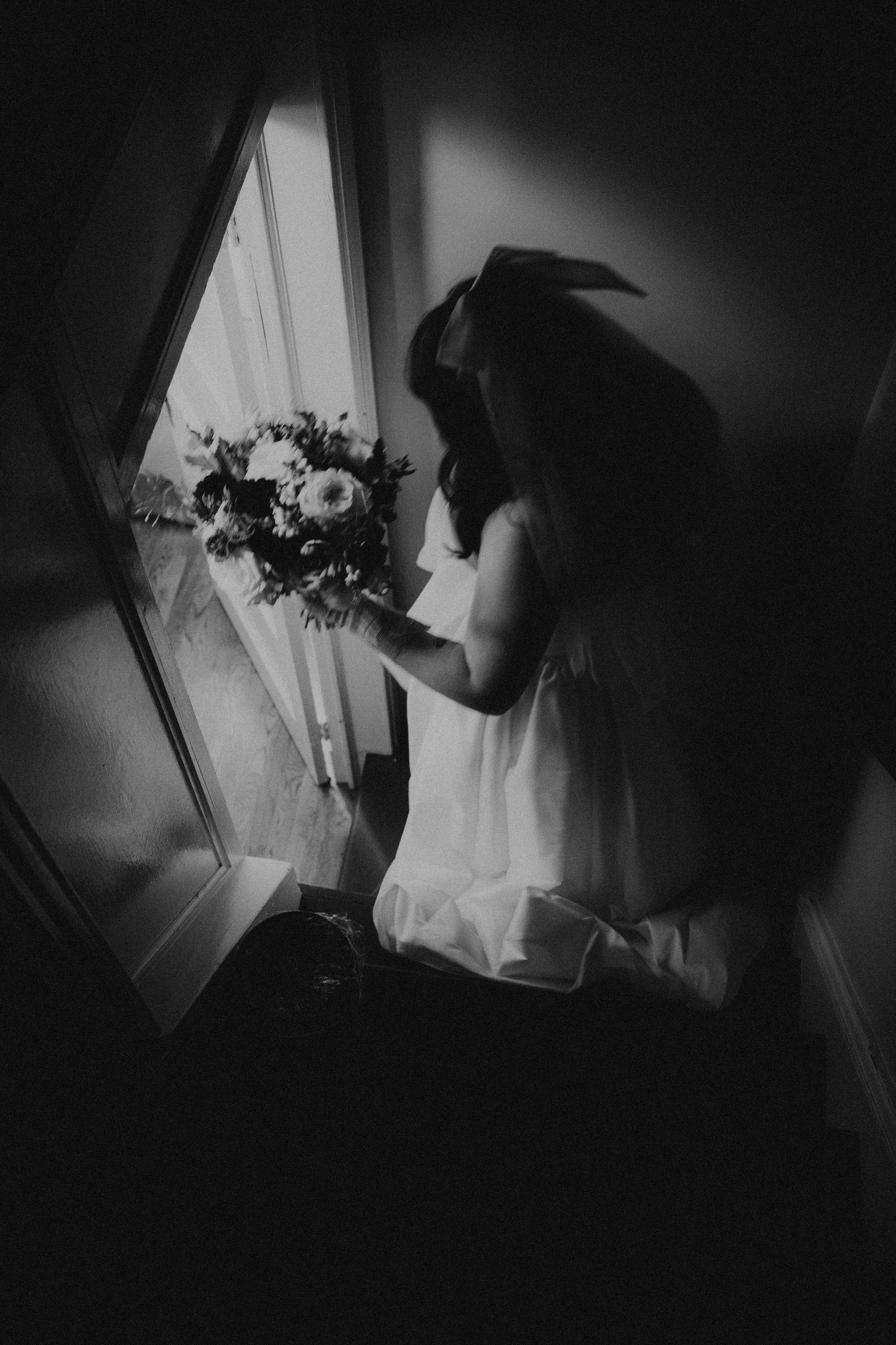 Black and white photo of a bride in a veil holding a bouquet while gazing out a window on her wedding day.