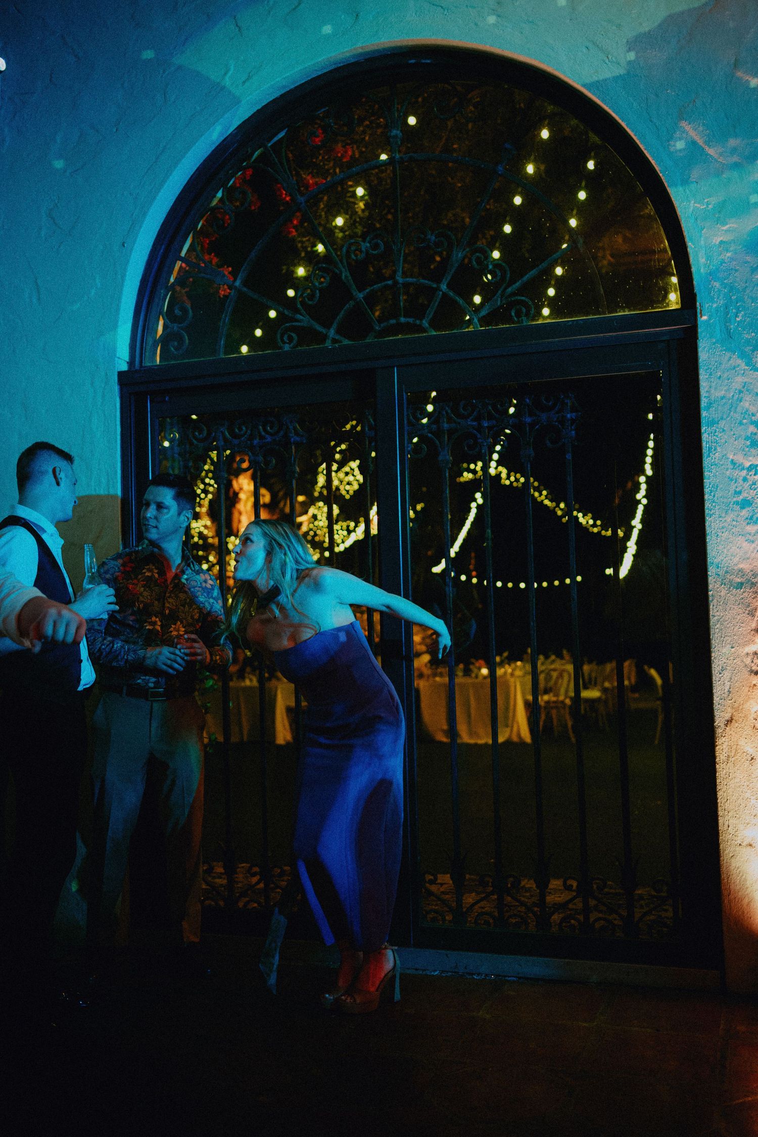 Woman in blue dress dancing near ornate iron gate with fairy lights at an elegant evening event.