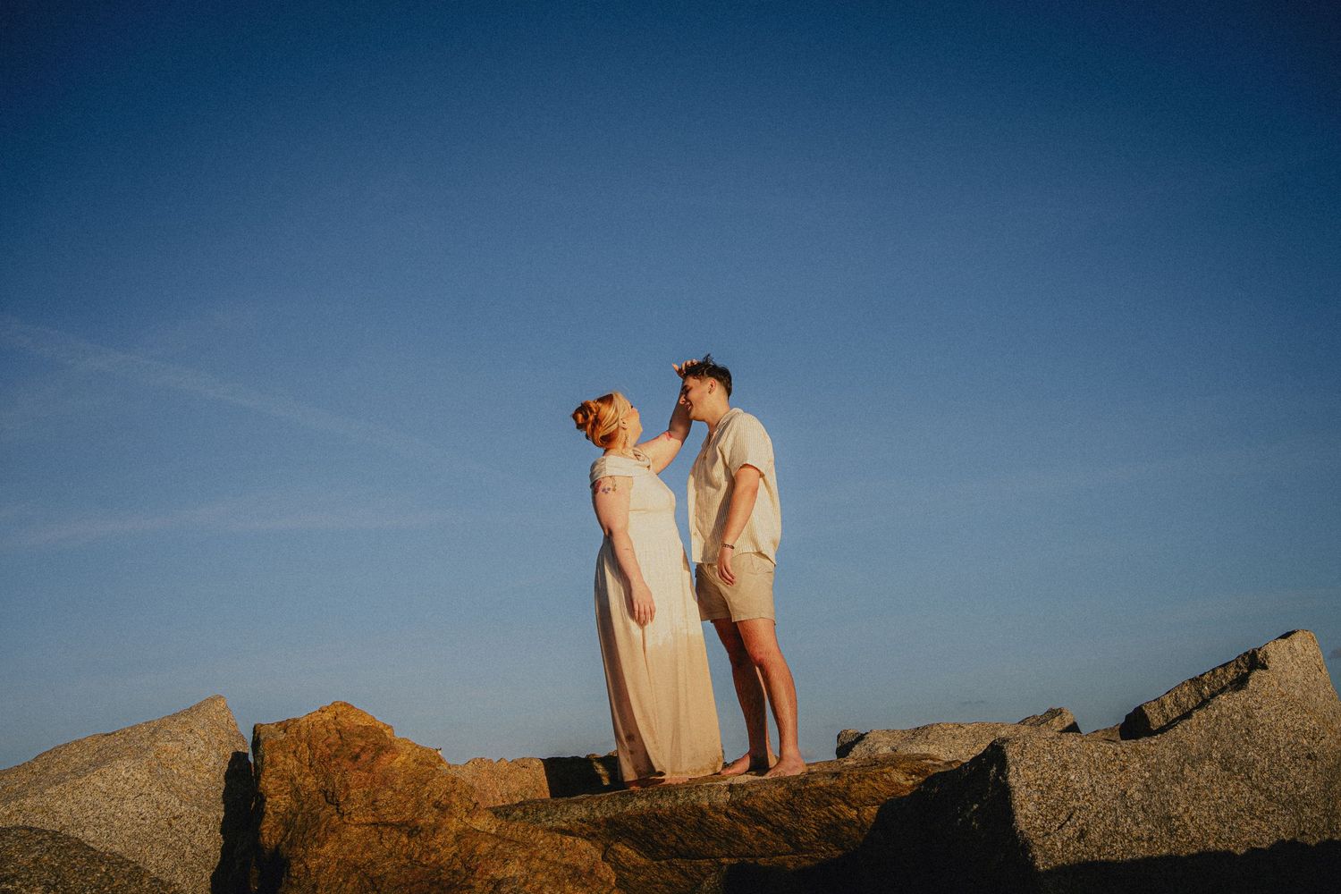 Couple standing on rocky shore at golden hour, woman in white dress, man in casual attire, blue sky backdrop.